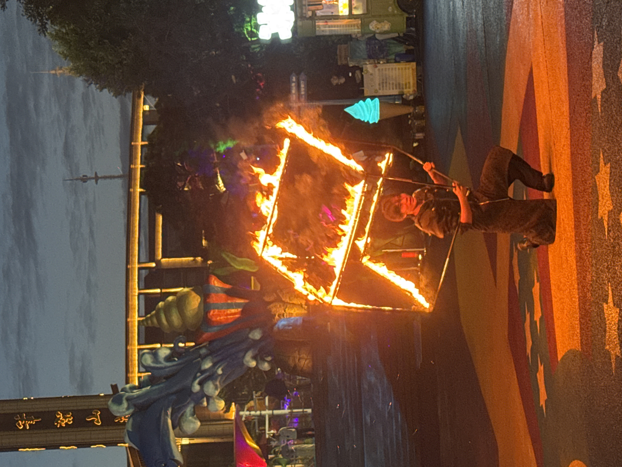 In the city of Taihe District, China, an outdoor performance is taking place at dusk. A performer, dressed in dark, loose-fitting attire, is kneeling on a colorful ground marking, holding a large, square object engulfed in flames. The flames are bright orange and yellow, with wisps of smoke rising into the air. The performer appears to be controlling the fiery object with two long sticks.
In the background, a large, elaborate structure with blue, wave-like elements and a yellow, spiral-shaped top is visible. Behind this, a brightly lit walkway or bridge with yellow lights stretches across the scene. Further in the distance, silhouetted trees and two tall towers with antennas are discernible against the twilight sky. To the right, a green food cart with a glowing blue ice cream cone and a menu board is situated. The overall atmosphere suggests a lively entertainment venue or festival.