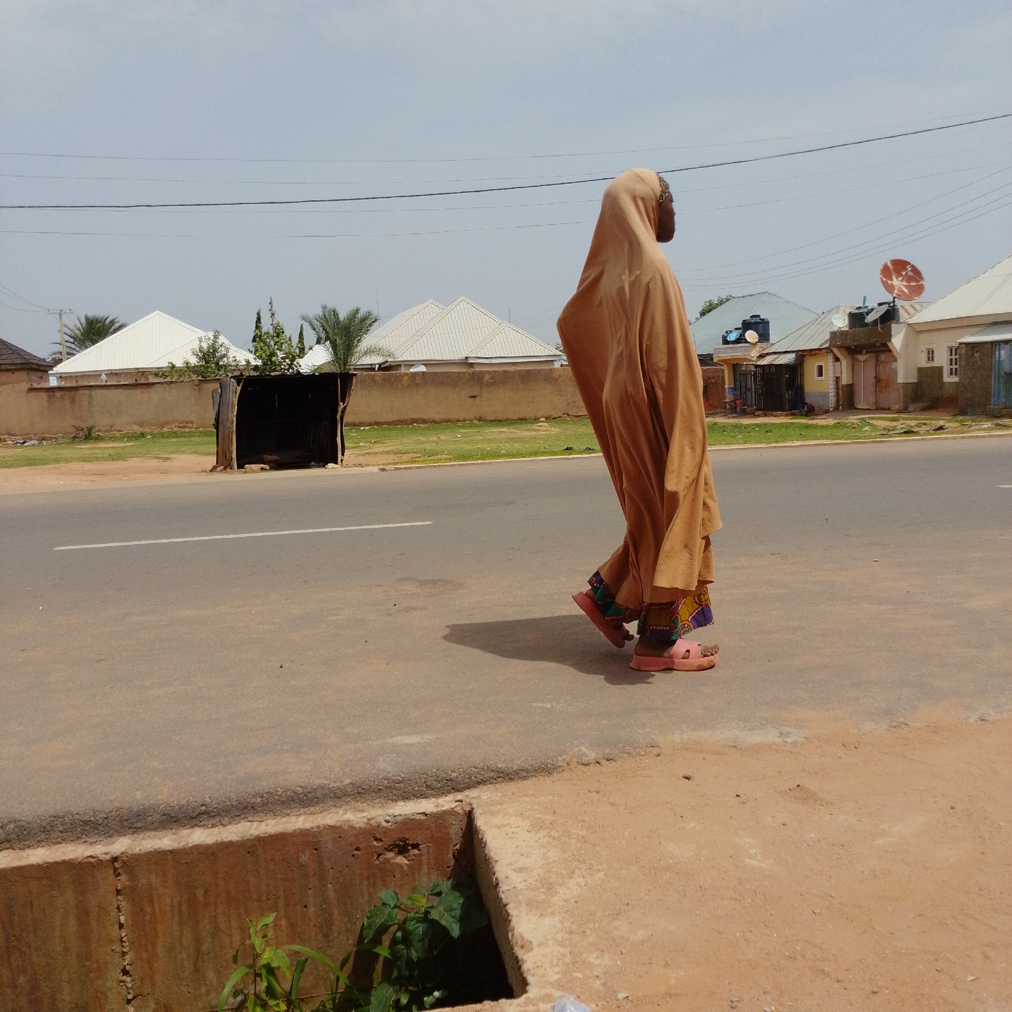 The image depicts a lone individual wearing a full-body tan garment, including a headscarf, walking on the side of a paved road in Gombe, Nigeria. The person is wearing pink sandals and appears to be moving away from the camera. The environment includes a roadside ditch with some vegetation, a light brown wall, and several buildings with corrugated metal roofs in the background, some of which have satellite dishes. Power lines are visible against the sky, which is overcast. A dark, open-sided structure resembling a makeshift shelter is located near the wall.