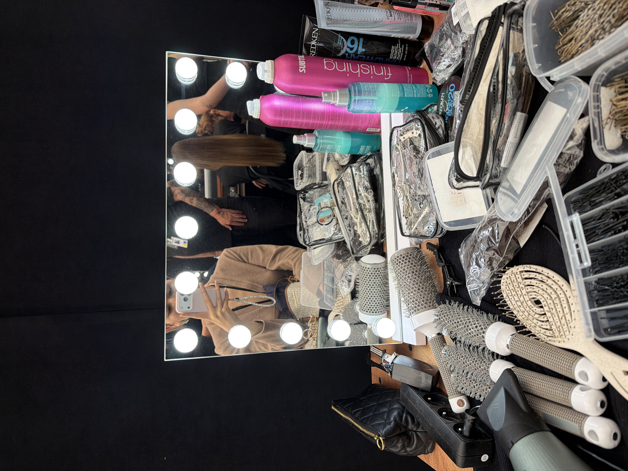 A mirror with lights is on a black surface. The reflection in the mirror shows a woman taking a photo. Several hair styling tools are visible, including brushes, hair spray, and hair clips. The location appears to be a dressing room or salon. There is no information given in this image to determine that it is in Cogolin, France.