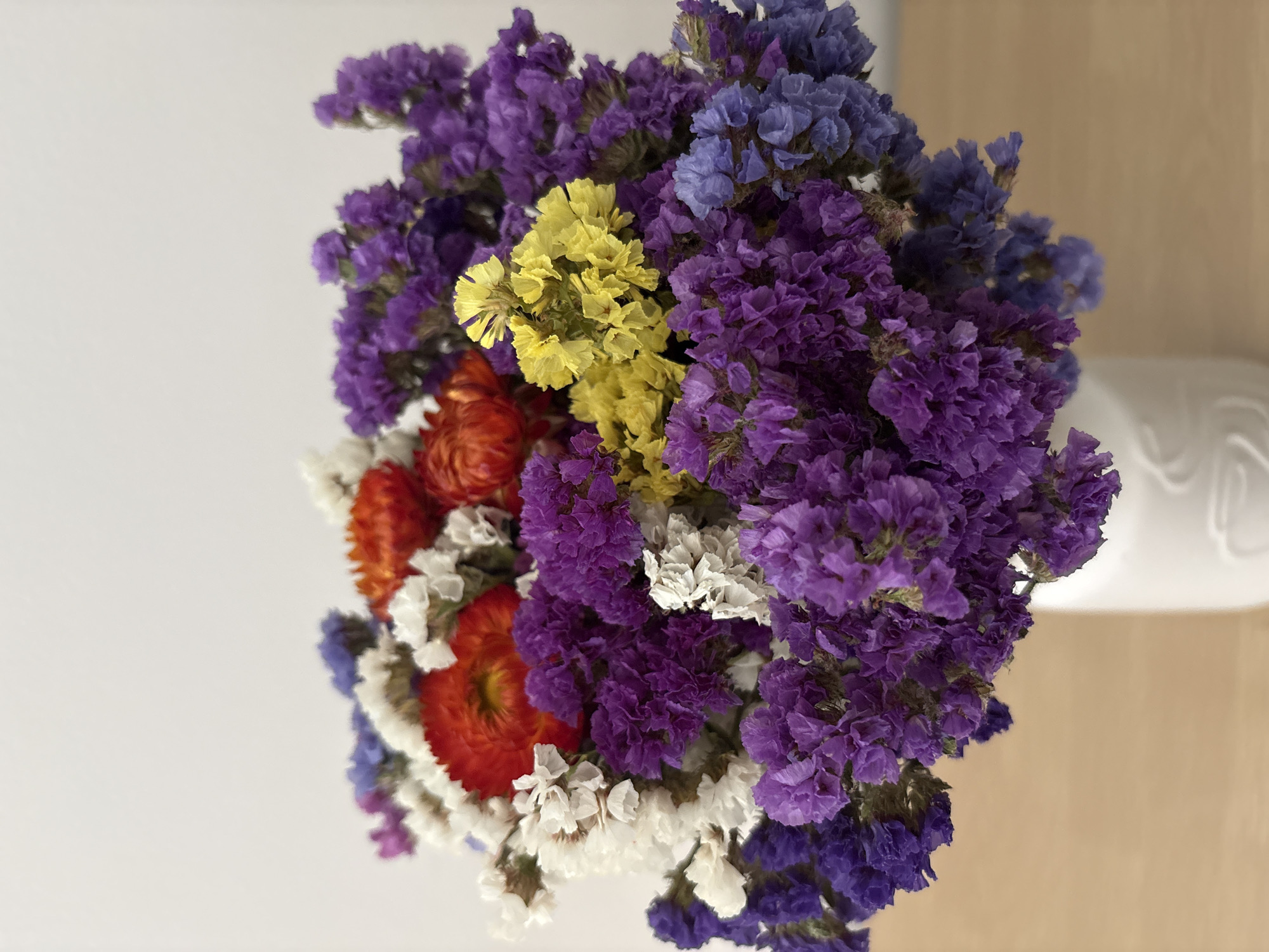 A vertical close-up shot displays a vibrant bouquet of dried flowers in a white ceramic vase, set against a plain, light-colored background. The flowers are predominantly shades of purple, with clusters of smaller blooms in white, red, orange, and yellow interspersed throughout. The bouquet is full and arranged in a rounded shape. The vase has a subtle embossed pattern. The surface beneath the vase appears to be a light brown wooden table. The scene is well-lit, likely with natural light, casting soft shadows. There are no people present. This image is not associated with the city of Liberec, Czechia.