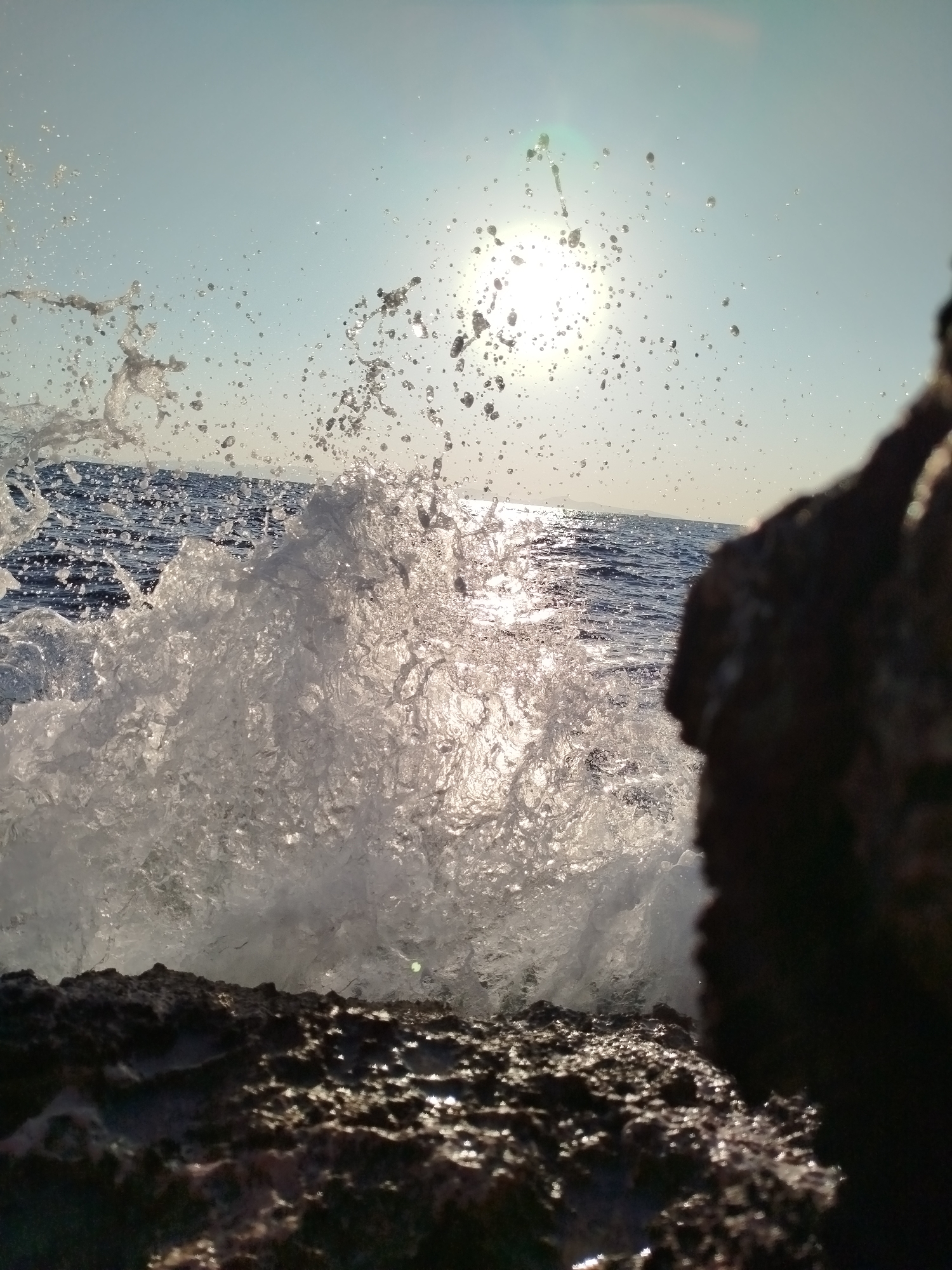 An extreme close-up captures a dynamic wave crashing against dark, wet rocks in Karaburun, Turkey. The sun, high in the sky, shines brightly, creating a lens flare and illuminating the spray of water droplets as they erupt from the impact. The ocean's surface is visible in the background, with a slightly hazy horizon. A large, dark rock formation dominates the right side of the frame, partially obscuring the view of the sea. The foreground is composed of rough, textured rocks, reflecting the sunlight. The image appears to have been taken during daylight hours, with the sun at its zenith. There are no people or animals visible in the scene.