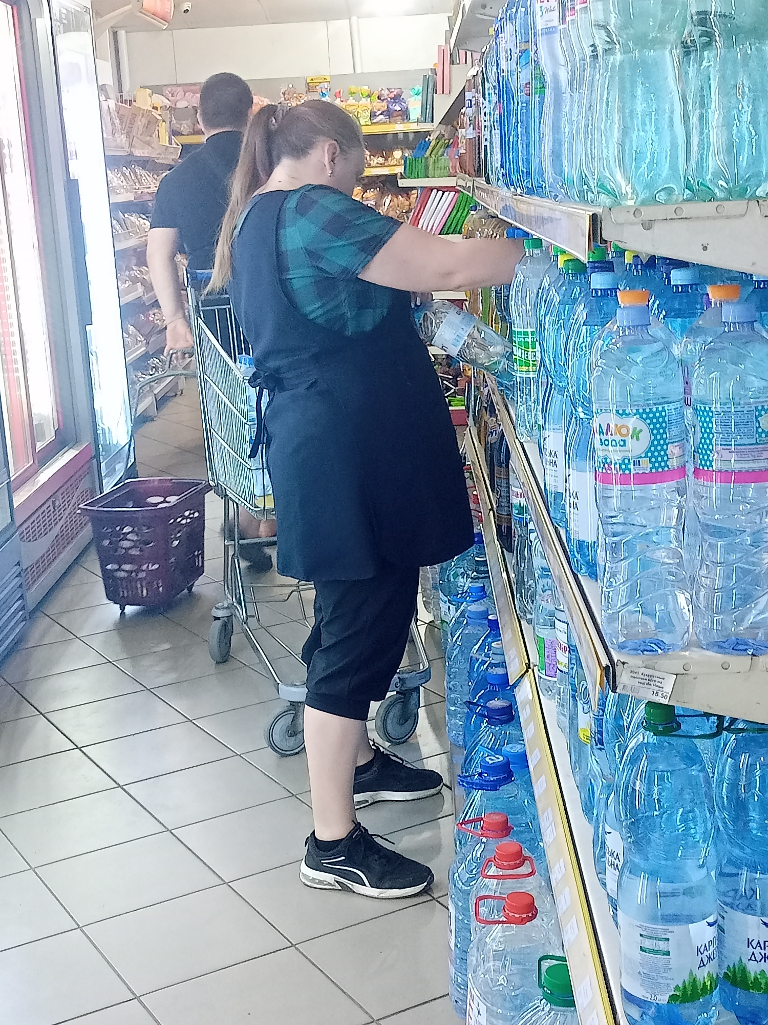 In a supermarket aisle in Bilhorod-Dnistrovskyi, Ukraine, a woman with her hair in a ponytail, wearing a dark green plaid shirt and a black apron over black pants, is reaching for a bottle of water from a shelf. She is standing next to a shopping cart. To her left, a man wearing a black t-shirt is partially visible. The shelves are stocked with numerous bottles of blue liquid, likely water. The floor is tiled with a grid pattern. To the left of the aisle, a red shopping basket is on the floor.