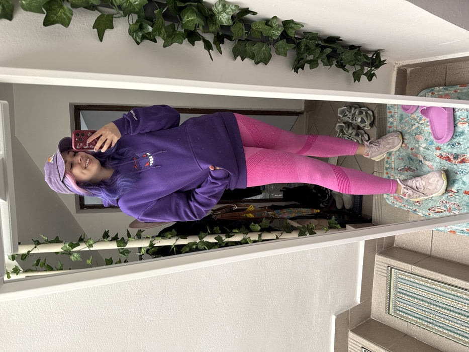 A young adult female is depicted in a full-body mirror selfie, standing indoors within a residential or entryway setting in the City of Zagreb, Croatia. She is smiling, looking towards her phone, which she holds in her right hand, obscuring part of her face. Her left hand is partially inserted into the front pouch pocket of her purple hooded sweatshirt, which features a graphic of the Pokémon character Gengar. She wears a faded purple baseball cap and pink athletic leggings with a textured pattern. Her hair, visible beneath the cap, is dyed purple with pink highlights. She wears light pink sneakers on her feet.
The environment captured in the mirror reflection includes a light-colored wall with an artificial green ivy vine decoration running vertically along its right side. The floor beneath her is covered by a rectangular rug with a light blue and multi-colored butterfly pattern. To her right, a pair of gray sneakers and a single pink slide sandal are visible on the floor. In the background, beyond the woman, is a doorway and what appears to be a coat rack or stand with various items hanging, including a multi-colored umbrella. To the left of the mirror, a tiled staircase with a textured runner is visible, indicating an interior multi-level space. The lighting suggests an indoor setting, possibly with natural light from an unseen source or overhead artificial lighting.