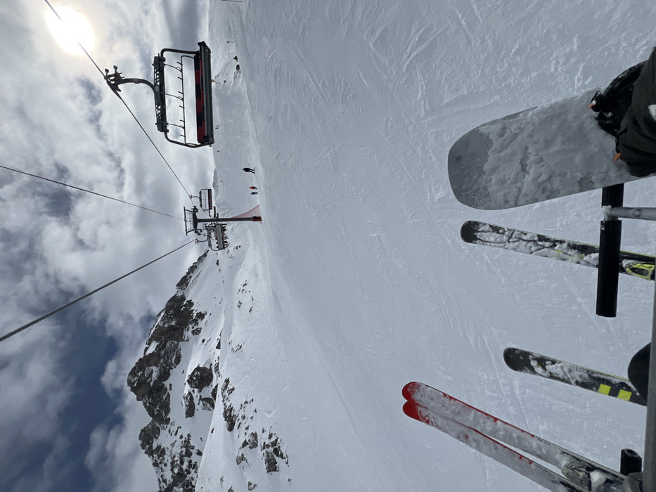 The original image, a rotated vertical shot, presents a first-person perspective from a ski lift ascending a snowy mountain, characteristic of a ski resort like Val-d'Isère, France.
In the immediate foreground, the skis of the person taking the photo are visible, secured to the footrest of the ski lift chair. One pair of skis is predominantly red, while another pair shows yellow and black accents. A snowboard is also prominently featured, attached to the same footrest.
Below and stretching into the distance, a wide, snow-covered ski slope is visible, marked with numerous faint tracks from previous skiers. Several small figures, identified as skiers or snowboarders, can be seen on the slope. Further down the mountain, another ski lift chair, occupied by multiple passengers, is seen approaching a red-accented support pylon.
To the left, a rugged, snow-dusted rocky mountain peak rises under a partly cloudy sky. The bright sun is visible in the upper left corner, creating a strong glare through the clouds. The scene captures an active daytime environment at a ski resort, with conditions appearing bright despite the cloud cover. There is no discernible text visible in the image.
