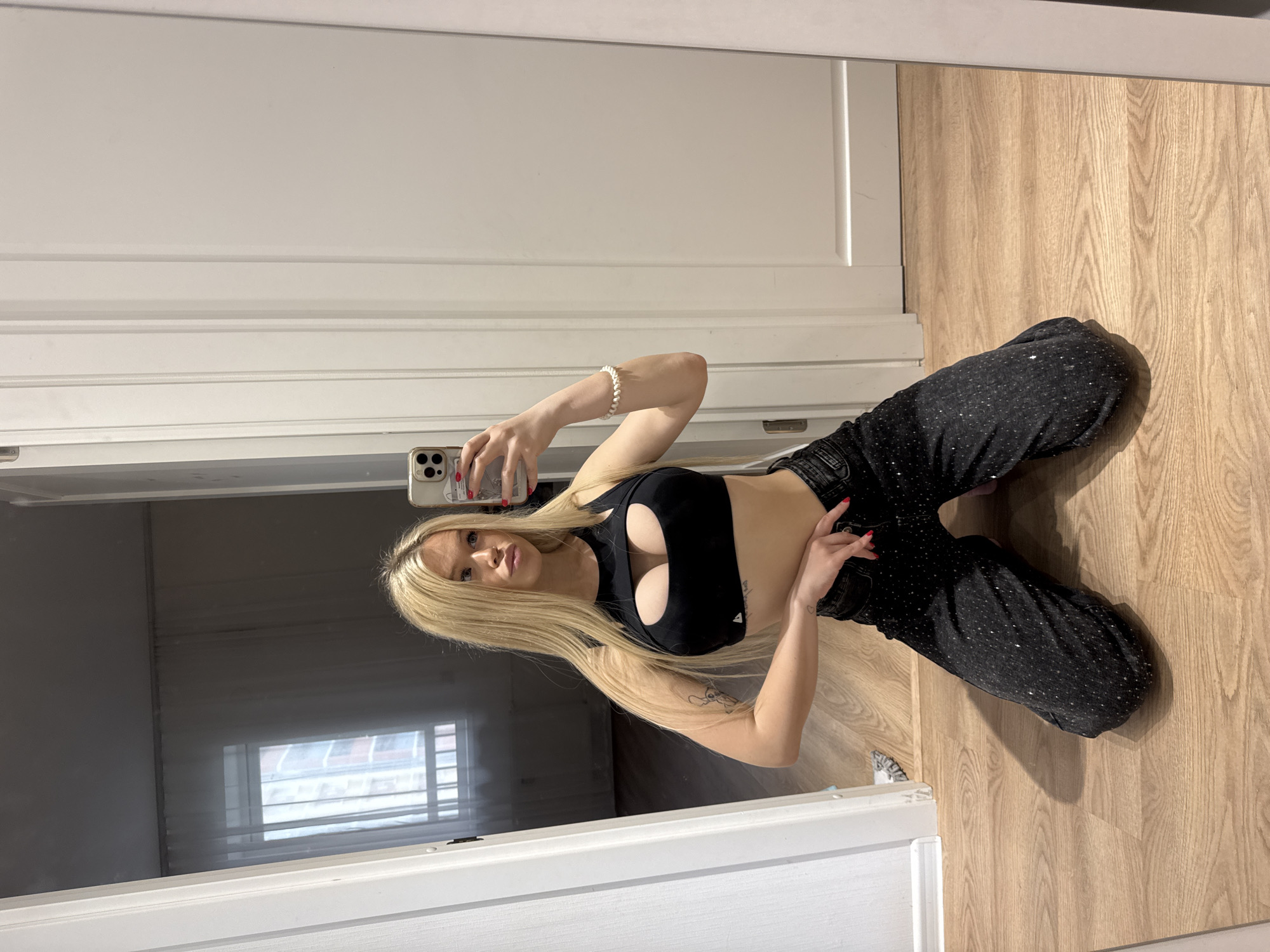 The media file shows a young woman with long blonde hair taking a mirror selfie in what appears to be an indoor setting, possibly a room or hallway. She is wearing a black crop top that exposes her midriff and has a cut-out detail over her chest, and dark, sparkly pants. The woman is sitting on a wooden floor, leaning back against a white door or wall. Her left arm is bent, and she is holding a smartphone in her hand to take the picture. Her right hand is resting on her thigh, and she is looking towards the phone.
In the background, visible in the mirror, is another door or wall, and a window with horizontal blinds. The overall lighting suggests it could be daytime, but the exact time is not clear. The setting appears to be residential. The woman's expression is neutral. There is a tattoo visible on her left arm. A white beaded bracelet is on her right wrist. The floor is made of light-colored wood planks. The image is taken from a slightly elevated angle.