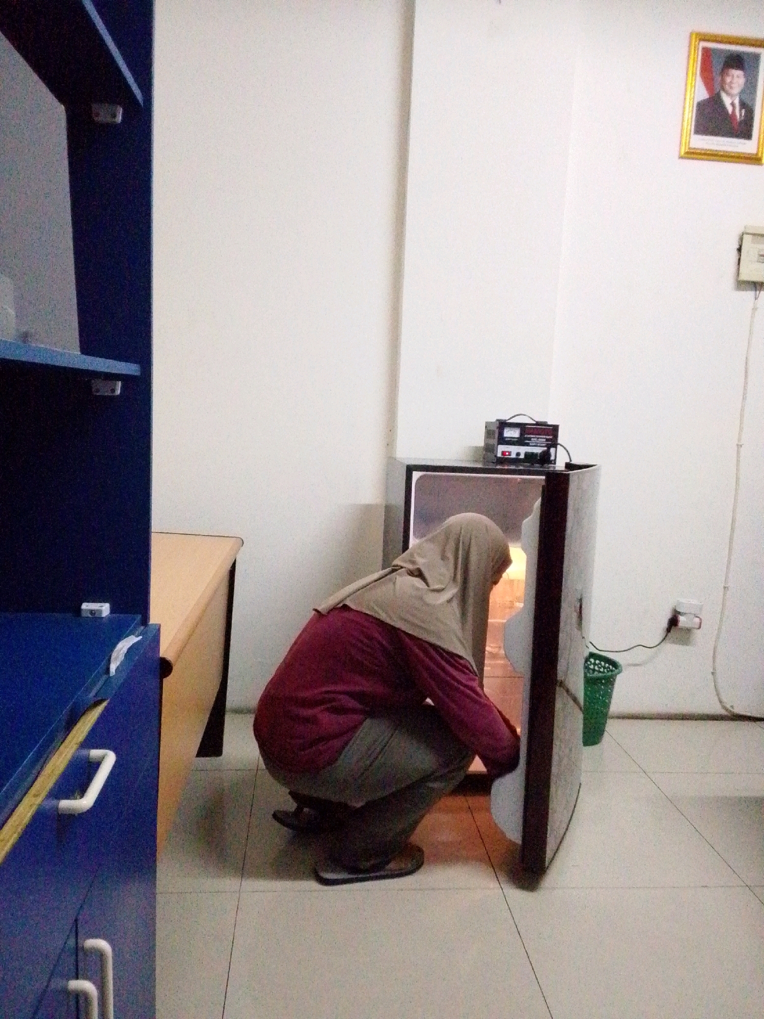 In a dimly lit room, a person wearing a hijab and a maroon shirt is squatting and reaching into an open, compact refrigerator. The refrigerator is located to the right of a blue cabinet with white handles. A tan desk is partially visible to the left of the refrigerator and behind the blue cabinet. On the wall above the refrigerator, a framed portrait of a man in a suit and hat is displayed, accompanied by the Indonesian flag. Electrical outlets and a green trash bin are also present on the wall near the refrigerator. The floor is tiled. The scene appears to be indoors, possibly an office or dormitory room in North Jakarta, Indonesia.