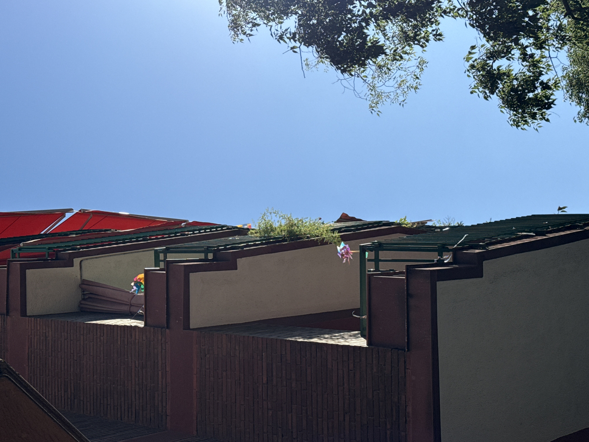 The image shows a view of a building's facade under a clear blue sky, with some tree foliage visible at the top of the frame. The building has a repeating pattern of balconies or terraces. The exterior walls are light beige or tan in color, and there are brown brick-like sections and dark green metal frameworks supporting the structures. Some balconies have objects like rolled-up mats and colorful decorations, possibly windmills. The light is bright, creating deep shadows. The location is likely in Barcelona, Spain, based on architectural style and potential context cues.