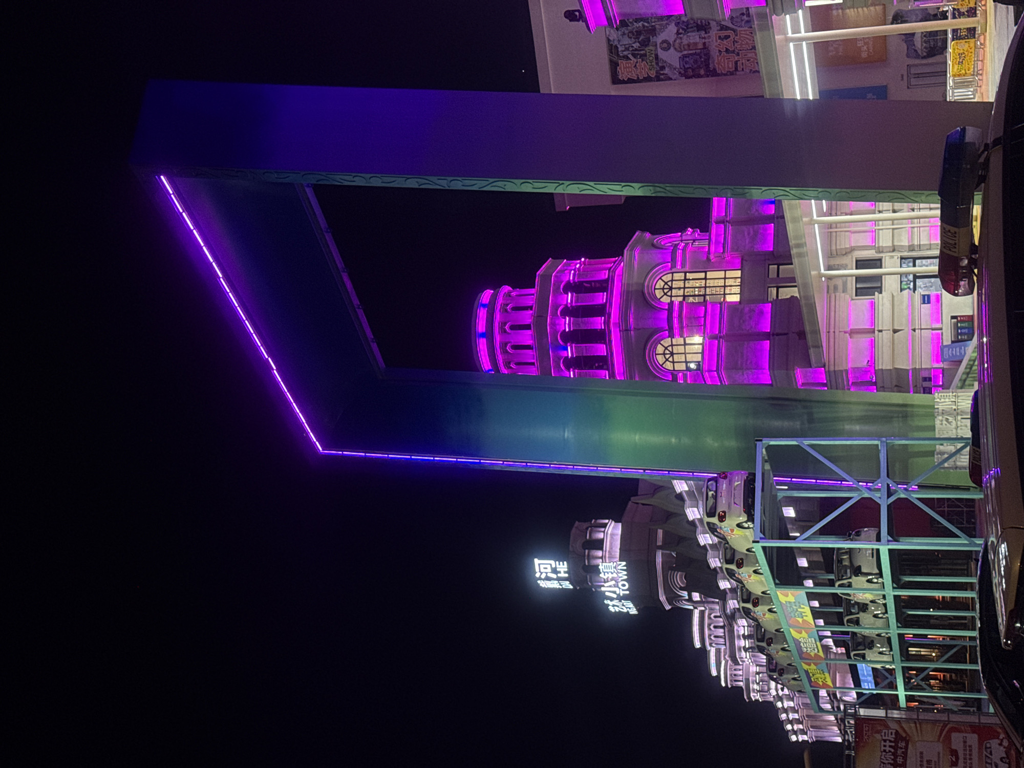 The image depicts a nighttime scene in the city of Qinhuangdao, China, likely in an entertainment or commercial district. Dominating the foreground is a large, illuminated structure with purple and green LED lights. To the left, a row of buildings with ornate architecture is visible, lit with vibrant pink and white lights. Several small cars are displayed on a metal rack in front of these buildings, illuminated by yellow and white lights. A police car with its lights activated is parked in the immediate foreground. The overall ambiance is festive and commercial, with a focus on bright, artificial lighting against the dark night sky.