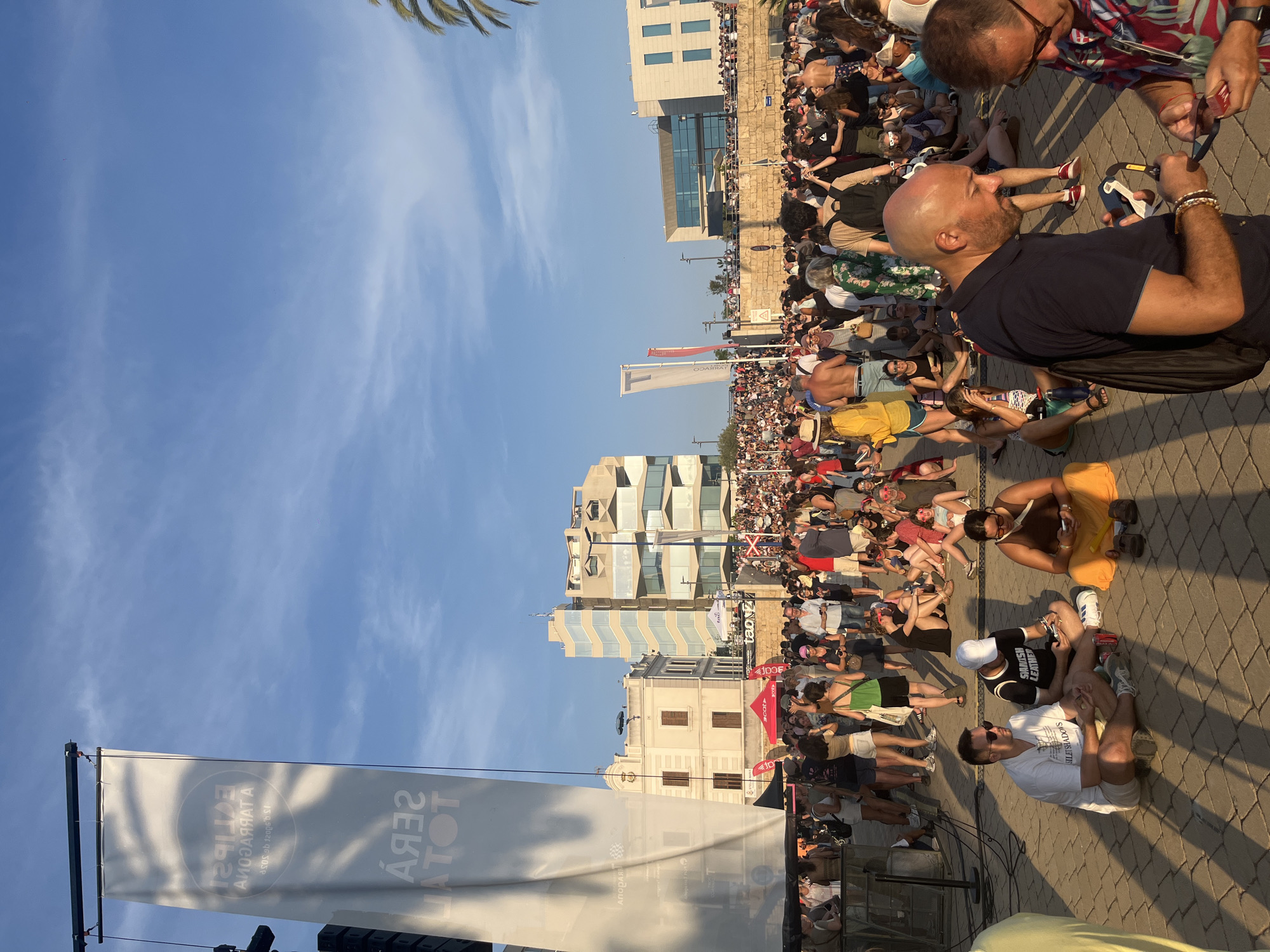 The image depicts a large outdoor gathering of people in the city of Tarragona, Spain, likely at a festival or event. The scene is dominated by a crowd spread across a paved area with a backdrop of modern buildings and a clear blue sky. A large white banner, partially visible on the left, displays text in what appears to be Catalan and possibly Spanish, including "SERA TOTAL" and "ECLIPSE TARRAGONA," suggesting the event is related to an eclipse and the city. In the foreground, several individuals are seated on the ground, some looking at their phones, while others engage in conversation. The crowd extends into the midground and background, with people standing and moving. The environment suggests a warm, sunny day, with shadows cast by the setting sun or overhead structures. The overall atmosphere appears to be relaxed and social, characteristic of an outdoor event.
