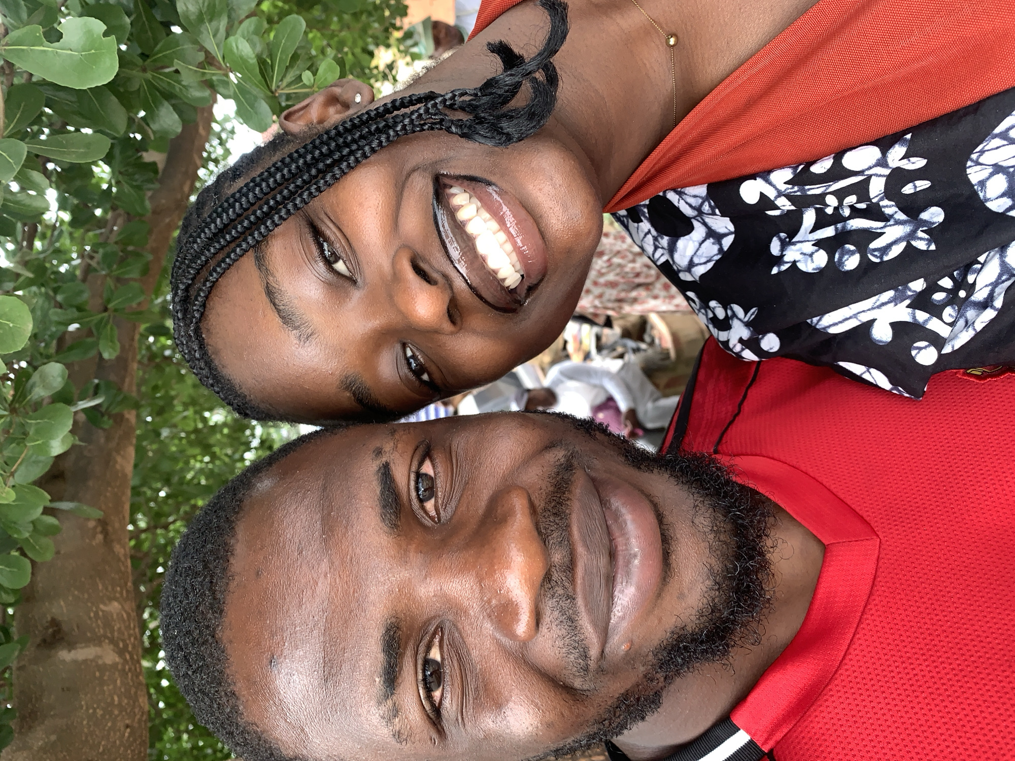 A close-up, eye-level, outdoor selfie shows a man and a woman in Rafin Par, Nigeria, with a tree canopy overhead. The man, on the left, has dark skin, a neatly trimmed beard, and is wearing a red jersey with black and white stripes on the collar. The woman, on the right, has dark skin and her hair is styled in braids that fall over her shoulders. She is wearing a black and white patterned garment over an orange top, with a delicate gold necklace. Both individuals are smiling broadly at the camera, with the woman's teeth gleaming due to lip gloss. In the background, out of focus, are other people and objects, suggesting a public or social gathering. The lighting indicates it is daytime.