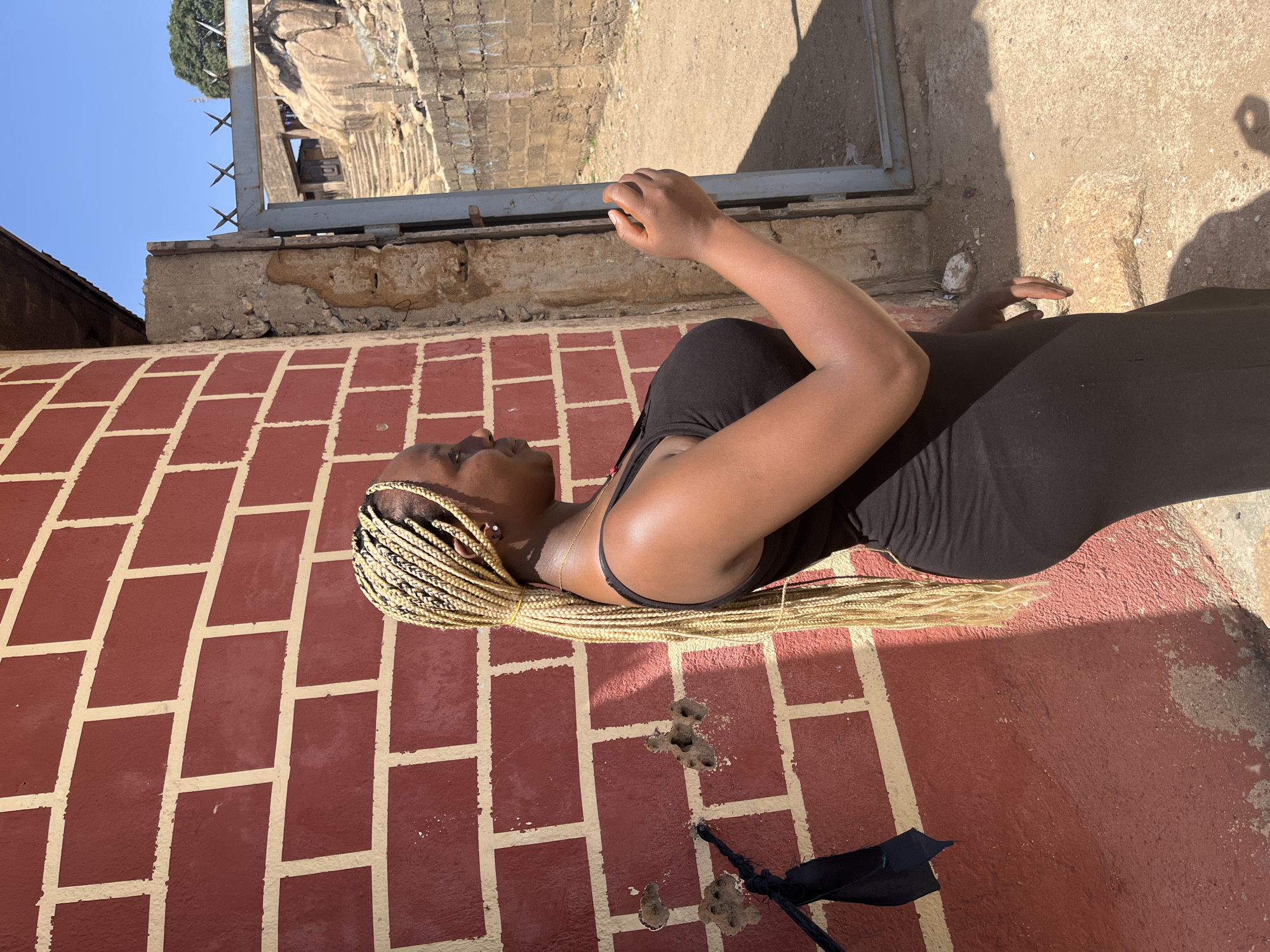 The image shows a person lying on a brick surface with a dark dress and blonde braided hair. The brick surface is painted in a red and tan pattern and has some damage near the person's head. The person appears to be holding a cell phone. There is a metal frame in the background that shows a view of a rocky hill. The location is Unknown, Unknown.
