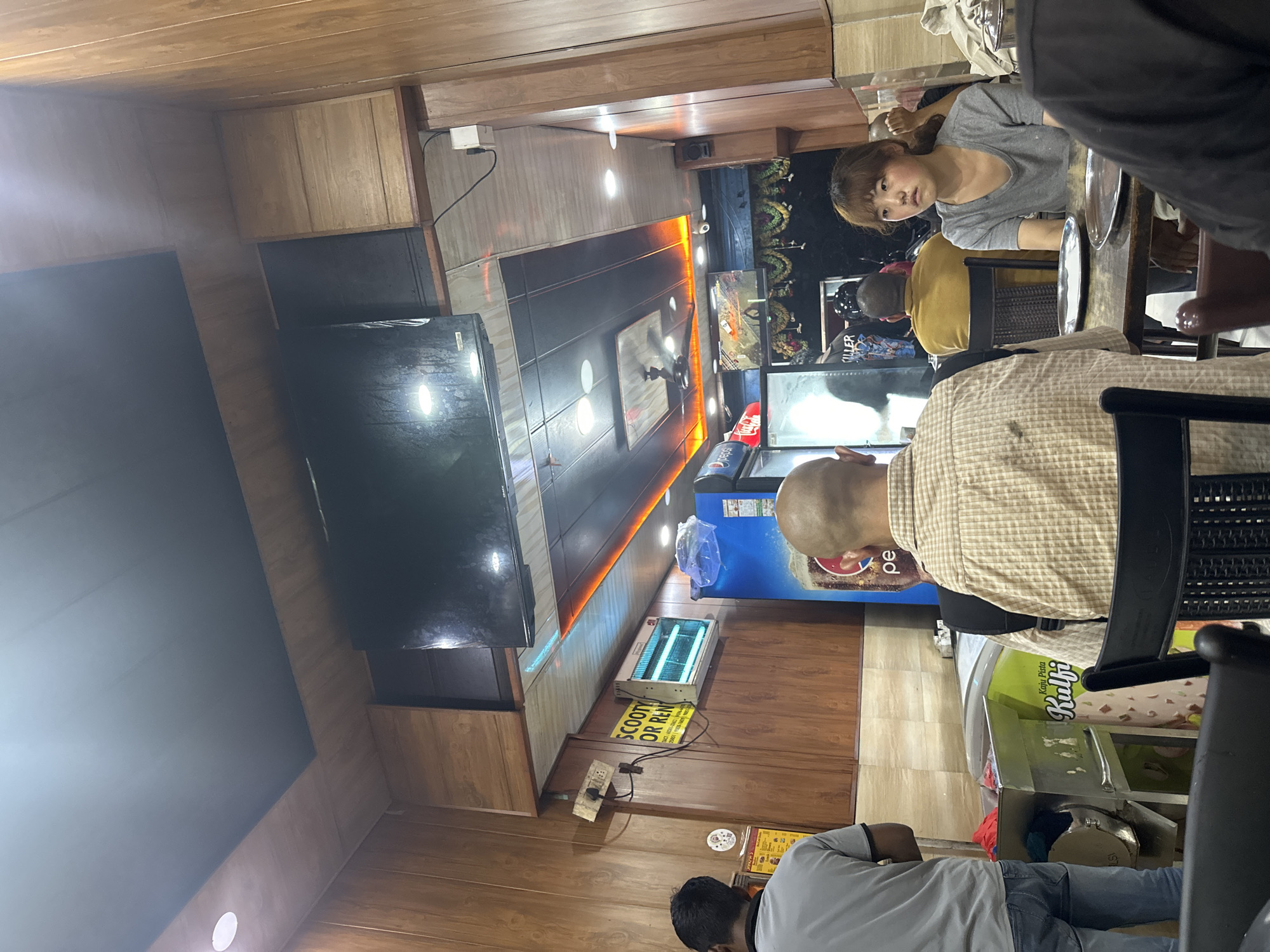 The image depicts the interior of a casual dining establishment, likely a restaurant or eatery, in an unknown location. The decor is characterized by wood-paneled walls and a suspended ceiling with wooden accents and orange LED strip lighting. A large flat-screen television is mounted on the ceiling.
In the foreground, a person wearing a grey t-shirt and blue jeans is partially visible on the left, facing away from the camera. To their right, a bald man in a checkered beige shirt is seated at a table, also facing away. In the middle ground, a young woman with dark hair and bangs, wearing a grey t-shirt, sits at a table looking towards the camera with a neutral expression. A Pepsi refrigerator is visible on the right side of the image. A sign advertising "Kaju Pista Kulfi" is affixed to a stainless steel counter.
Several other individuals are present in the background, their faces and activities largely obscured. The environment appears to be a functional and somewhat dated interior, likely a place of commerce given the presence of seating, a television, and food-related equipment. The overall lighting is dim, with some areas illuminated by the ceiling lights and the television screen.
