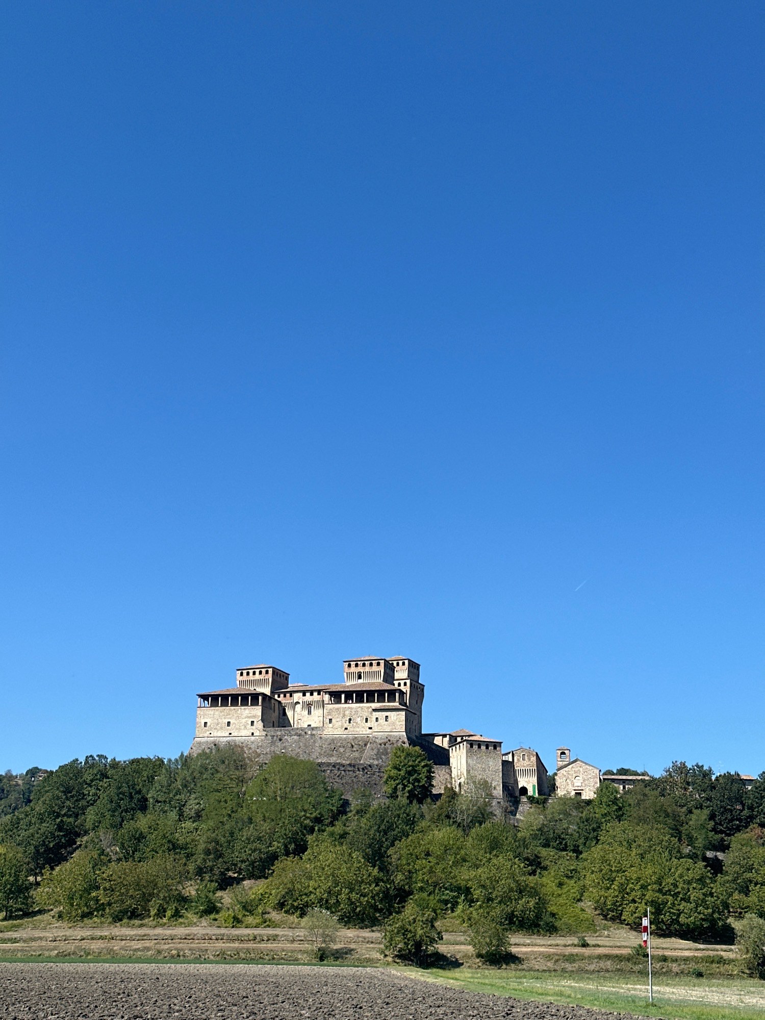 A daytime, eye-level, vertical shot captures the Castello di Torrechiara in Langhirano, Italy, situated on a hill. The castle, constructed of stone, features multiple towers and crenellations. It is surrounded by lush green trees, with terraced fields visible below. The sky is a clear, bright blue. In the foreground, a patch of tilled earth indicates agricultural activity. A flag is visible on the right side of the frame. No people are present in the image.
