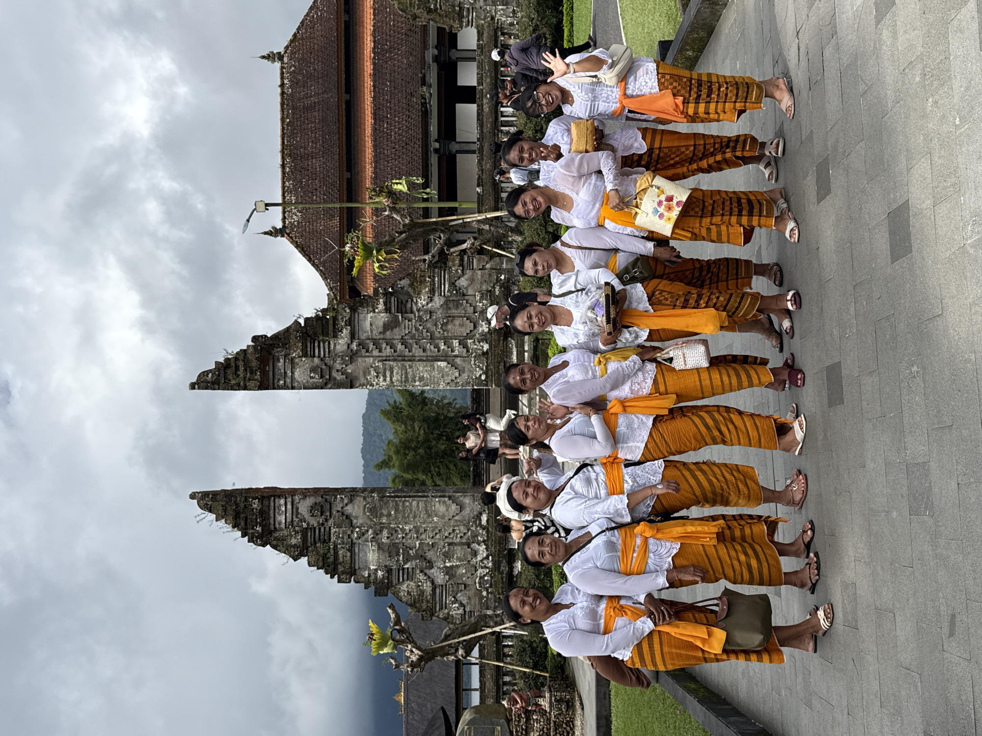 In Baturiti, Indonesia, a group of 11 women stand in a line on a paved surface in front of a stone temple structure. They are uniformly dressed in white blouses, orange sarongs, and white headbands. Some carry bags or clutches. The temple structure is ornate and weathered, with a tiered roof and sculpted stone elements. The sky is overcast. In the background, there are trees and a glimpse of hills or mountains. Some tourists are behind the main subjects.