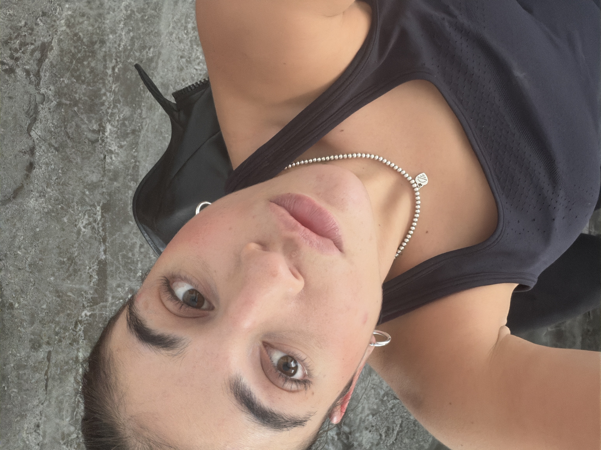 The image is a close-up, rotated portrait of a young adult female looking directly at the viewer. She has light-to-medium skin tone, dark brown eyes, and dark eyebrows. Her lips are slightly parted, revealing a neutral or subtly relaxed expression.
She is wearing a dark, possibly black, tank top with a mesh or textured pattern visible on the upper chest area. Around her neck, she wears a silver beaded necklace with a small, heart-shaped charm. The text on the charm is clearly visible and reads: "PLEASE RETURN TO TIFFANY & CO. NEW YORK". A simple silver hoop earring is visible in her right ear.
Behind her, a dark, possibly black, garment or bag with a zipper is partially visible on her left shoulder and arm. The background consists of a textured, grey surface, resembling concrete, stone, or a mottled wall, providing a plain setting for the portrait.
There is no specific activity depicted other than posing for the photograph. The lighting appears soft and even, suggesting an indoor setting or an overcast day. While the location context is given as Puebla, Mexico, there are no specific visual cues within the image itself to confirm this location.