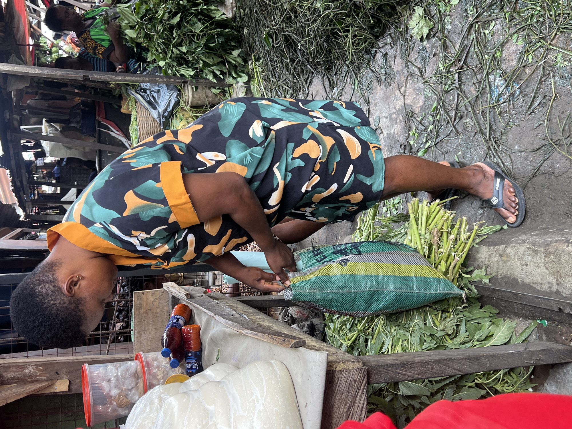 The image shows a woman in a busy outdoor market, likely Nkpor, Nigeria. She is wearing a colorful dress with a black background and a pattern of green, orange, and cream shapes. She is crouched down, tending to a large, green and yellow striped bag filled with produce, possibly vegetables or herbs. Her hands are busy working with the bag, suggesting she is either packing, unpacking, or arranging the contents.
The ground around her is damp and muddy, with scattered green vegetation, indicating it might have rained recently or the area is prone to wetness. Behind her, a glimpse of another person, also dressed in a green patterned top, can be seen amongst piles of more greenery and market stalls. The overall scene suggests a vibrant and active marketplace environment, with goods being handled and sold.
The lighting is natural, suggesting it's daytime. No specific weather conditions are evident beyond the damp ground. The presence of stacked produce, the woman's focused activity, and the background hints of other people and stalls all contribute to the atmosphere of a bustling local market. The image captures a moment of everyday commerce in a Nigerian setting.