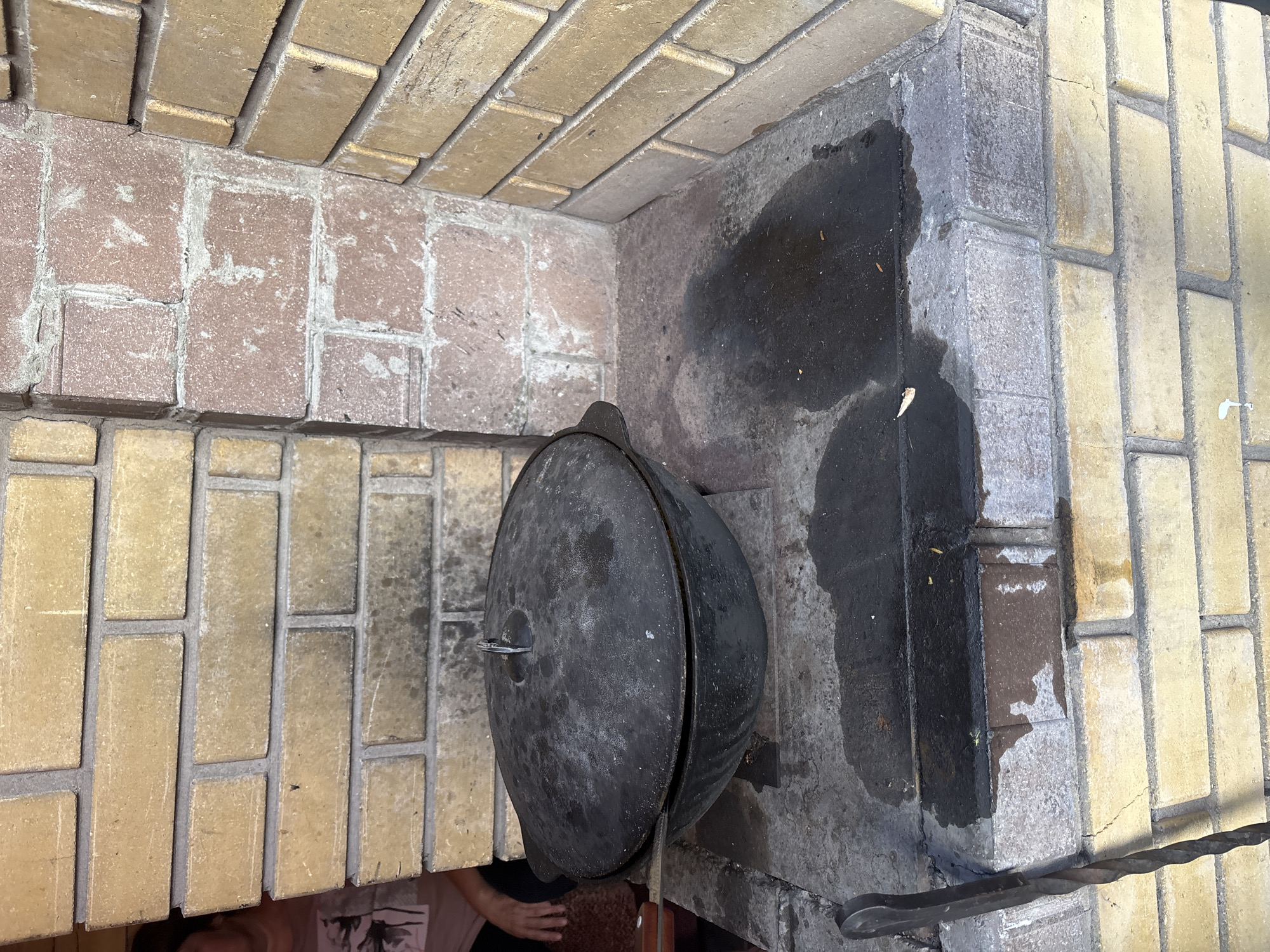 A large, black, cast-iron cauldron with a lid is placed in a brick oven. The cauldron is positioned to the left of the frame, with its lid slightly ajar, revealing a glimpse of its interior. The oven is constructed with yellow and pinkish-brown bricks, laid in a staggered pattern. The interior of the oven shows signs of use, with dark, sooty patches on the brickwork.
A metal handle, likely for the cauldron, protrudes from the lid, and a wooden-handled tool, possibly a knife, is placed on the edge of the oven. To the right of the cauldron, a dark, wet patch is visible on the floor of the oven, and a twisted metal poker rests on the brickwork. The background is filled with more brickwork, creating a textured and rustic setting.
The lighting suggests it might be daytime, but the specific time is not clear. The overall scene evokes a sense of outdoor cooking, possibly in a traditional setting, given the presence of the brick oven and cast-iron cauldron. A small portion of a person's arm and shoulder wearing a t-shirt can be seen in the lower left corner of the image. No visible text is present in the image. The location is contextually identified as Chernogorsk, Russia.