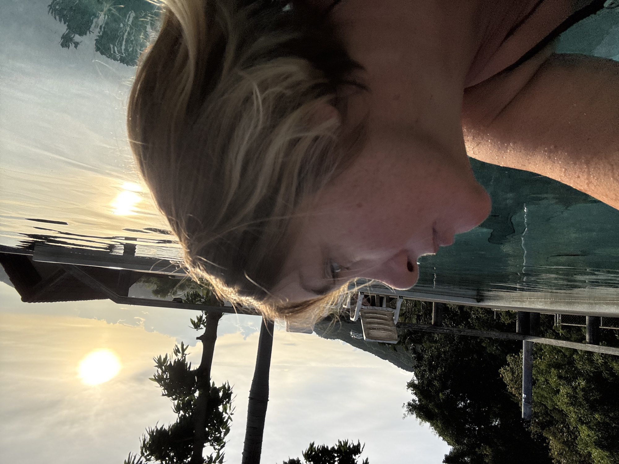 A woman with blonde hair is relaxing in a swimming pool at sunset. The water is calm, reflecting the golden light of the sun. In the background, there are lush green trees and rolling hills. The woman is looking up at the sky, with a serene expression on her face. The scene appears to be a tranquil oasis, possibly in a resort or private residence, likely located in the outskirts of Ban Bang Rak, Thailand, given the tropical vegetation and mountainous backdrop. There are lounge chairs by the pool, indicating a place of leisure. The overall atmosphere is one of peace and relaxation.