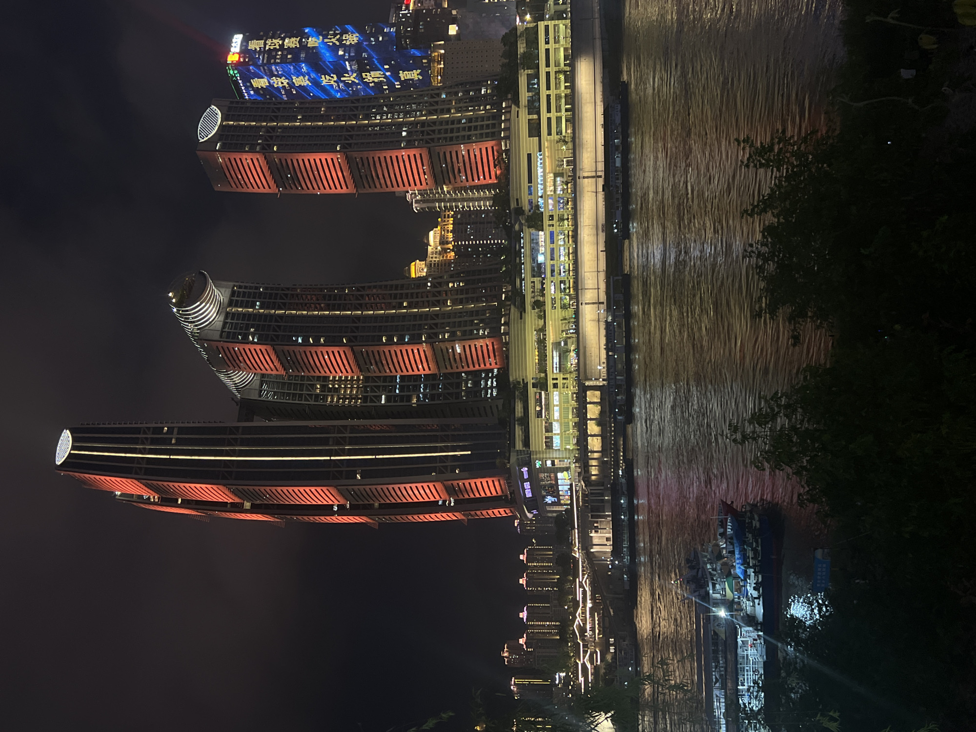 This nighttime photograph captures a vibrant urban waterfront scene in Jiangbei Cheng Subdistrict, China. The foreground features a wide river, its surface reflecting the colorful lights of the city. Several boats are docked or moving on the water, some appearing to be ferries or tour boats. Along the riverbank, a multi-story building complex is illuminated, with shops or restaurants visible on its lower levels. Above this, several striking modern skyscrapers dominate the skyline. The tallest and most prominent building on the left is illuminated with red and white vertical lights. To its right, another uniquely shaped skyscraper has a circular top section lit with white lights. Further to the right, a third skyscraper is adorned with bright blue lights forming Chinese characters. In the background, a dense cluster of other buildings, also lit up, extends into the distance. The sky is dark, indicating it is nighttime. The overall impression is one of a bustling and visually dynamic city at night, centered around its riverfront.