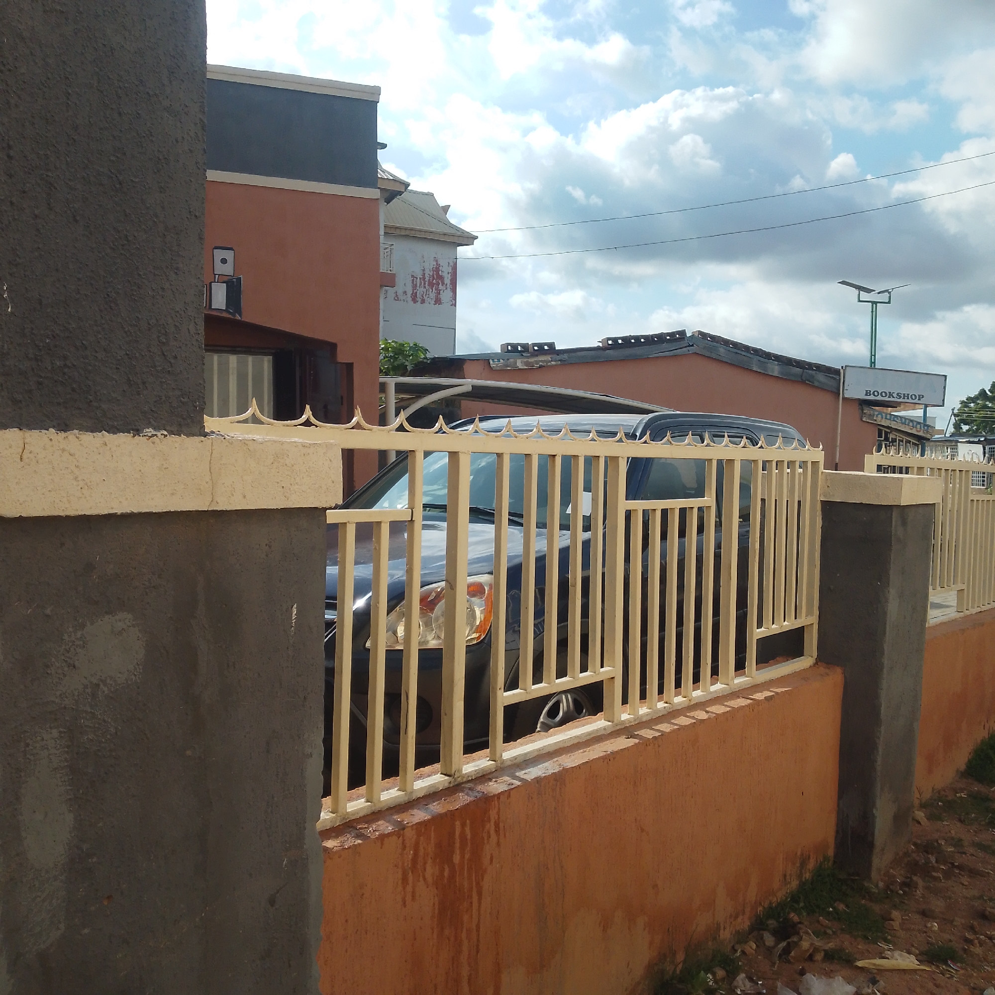The image captures a view of a street in Pantame, Nigeria, dominated by a cream-colored metal fence that runs across the foreground. Behind the fence, a dark-colored SUV is partially visible, parked on a property. The fence itself is made of vertical bars with decorative pointed finials at the top. The fence is mounted on a low, orange-colored wall that appears weathered and stained.
Beyond the fence and the SUV, several buildings are visible. To the left, a multi-story building exhibits a contrasting color scheme with sections of rust-orange and dark gray, topped by a cream-colored border. A portion of a dark-colored window or entrance is also visible on this building. To the right of this building, another structure with a light-colored roof and a white facade marked with red streaks is seen. Further to the right, a singular sign with the word "BOOKSHOP" in black lettering on a white rectangular background is mounted on an orange building. Beneath this sign, another sign with the same lettering is partially obscured. The sky above is partly cloudy, with white cumulus clouds scattered against a light blue background. A few power lines are visible stretching across the sky. The ground in the foreground consists of dirt with patches of dry grass and scattered debris.