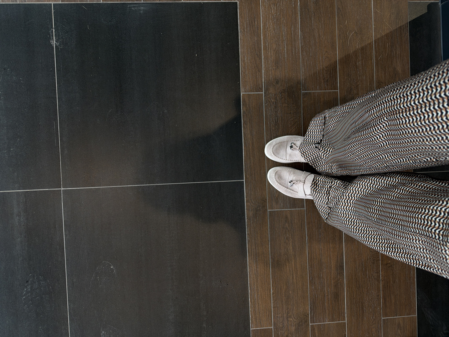An overhead shot captures a person's legs and feet standing on a tiled floor in Izhevsk, Russia. The person is wearing light gray loafer-style shoes and wide-leg trousers featuring a distinctive black, white, and brown wavy, zig-zag pattern.
The floor is distinctly divided into two sections. On the left side, large, dark gray or black square tiles are visible, with faint scuff marks and shoe prints scattered across their matte surface, suggesting regular foot traffic. On the right side, rectangular tiles designed to resemble medium-brown wood planks are laid in a staggered pattern. The clear transition between these two contrasting flooring types suggests a specific design choice within an indoor space, possibly marking a boundary between different functional areas like a hallway, an entrance, or distinct zones within a room.
The activity depicted is simply standing still. The lighting appears even and consistent with an indoor setting, likely under artificial light or diffused natural light. There are no discernible emotions conveyed, and no visible text is present in the image.