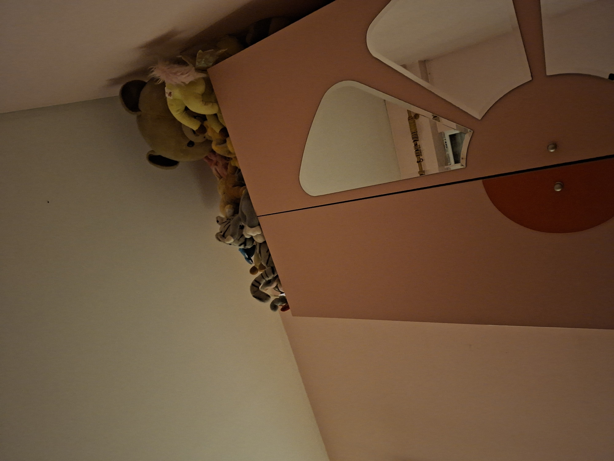 A low-angle, medium shot captures a corner of a room with a pink wall and ceiling. On top of a pink closet, a collection of plush toys are visible, including a large yellow teddy bear, a smaller yellow stuffed animal with pink hair, and other assorted stuffed animals. The closet doors have decorative, irregularly shaped mirrors. The overall impression is one of a child's room or a storage area for toys. The image does not provide specific details to determine a location within Pleven, Bulgaria.