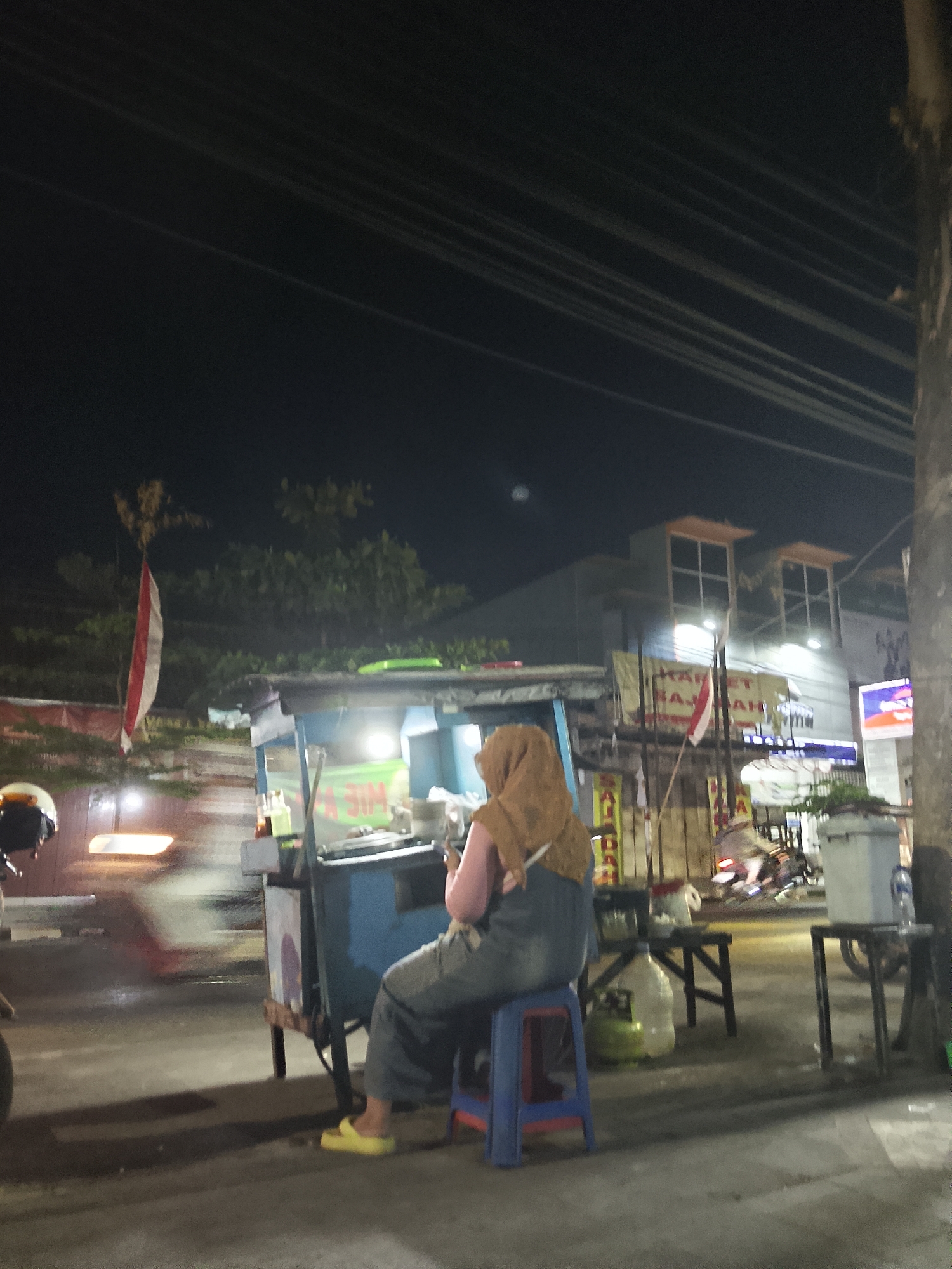 In Balun, Indonesia, a person wearing a headscarf and denim overalls sits on a blue stool next to a food cart. The cart is equipped with a light that illuminates the workspace, and various bottles and containers are visible. In the background, a building with distinctive architectural features, possibly featuring windows and orange accents, stands under a dark night sky. A red and white flag is displayed prominently, suggesting a national or local observance. The street is paved and appears to be wet in places, reflecting the artificial lights of the scene. Motion blur indicates the presence of moving vehicles, contributing to the dynamic atmosphere of the urban night.