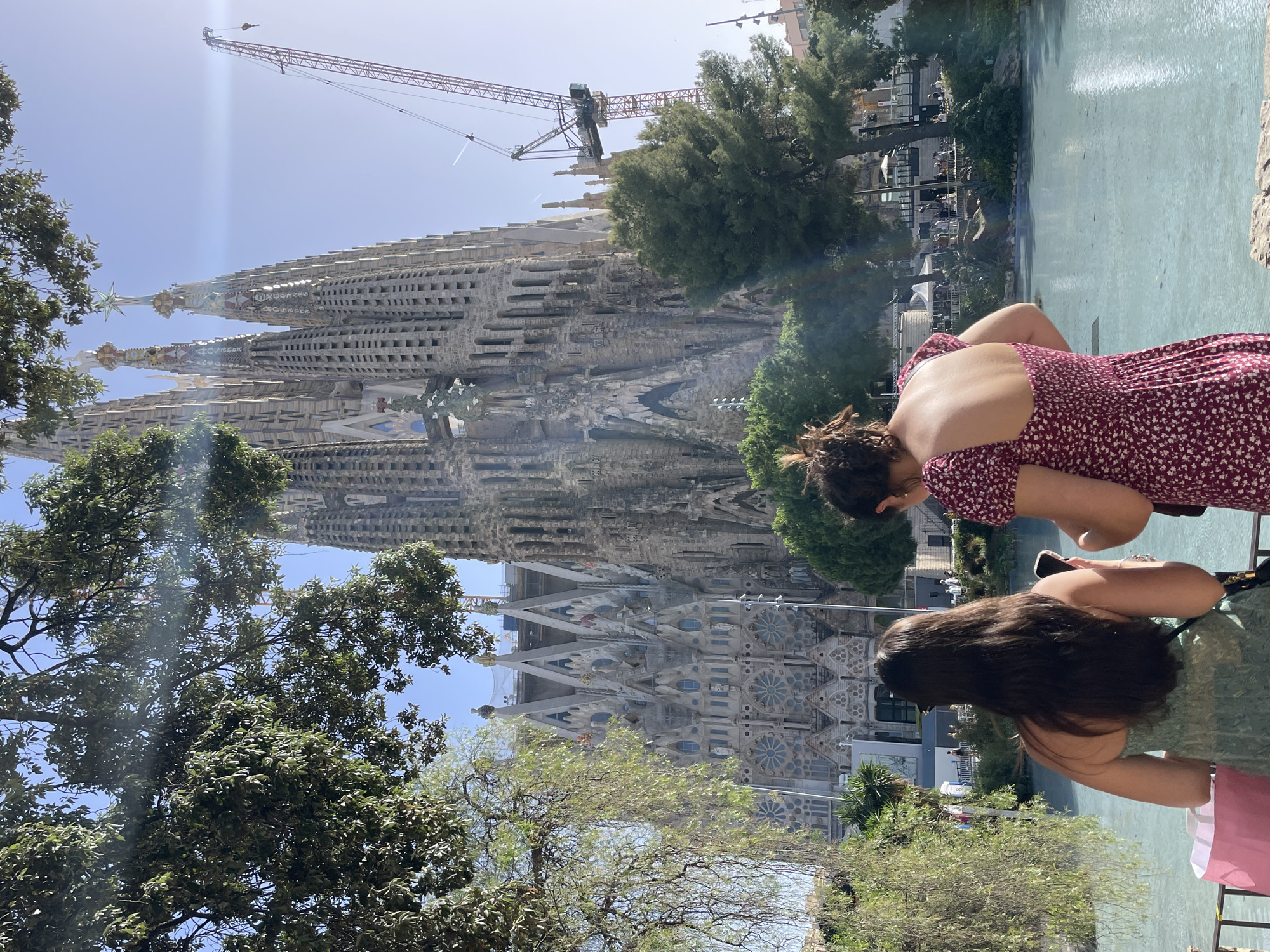 The image shows two women from behind, looking at the Sagrada Familia in Barcelona, Spain. The Sagrada Familia is a large, unfinished Roman Catholic minor basilica designed by Catalan architect Antoni Gaudí. In the background, a large construction crane is visible, indicating ongoing work on the basilica. The women are standing near a body of water, possibly a pond or pool, with trees and greenery surrounding them. The sky is clear and blue, suggesting it is a sunny day. The woman on the left is wearing a green dress and has long, dark hair. The woman on the right is wearing a floral print dress and her hair is in a bun. Both women are holding smartphones. A pink shopping bag is visible on the ground near the woman on the left.