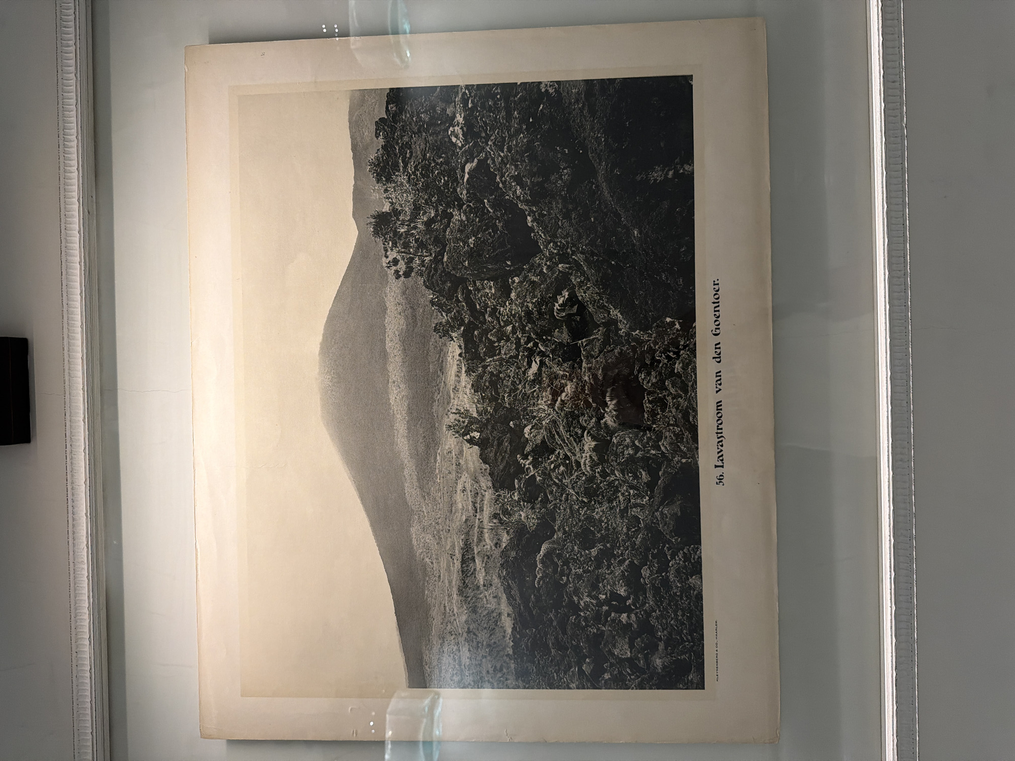 The image is a framed, black and white photograph or lithograph titled "56. Lavastroom van den Goentoer." It depicts a landscape with a prominent, light-colored mountain or hill to the left, contrasted by a dark, heavily textured area of what appears to be a lava flow on the right. The lava flow area has dense vegetation and intricate rocky formations. The scene is likely in a volcanic region, possibly near Mount Guntur (Goentoer) in Indonesia. The print has visible aging and is framed with a light-colored, ornate frame. The publisher is listed as N.J. KLETHEBERG & CO., HAARLEM.