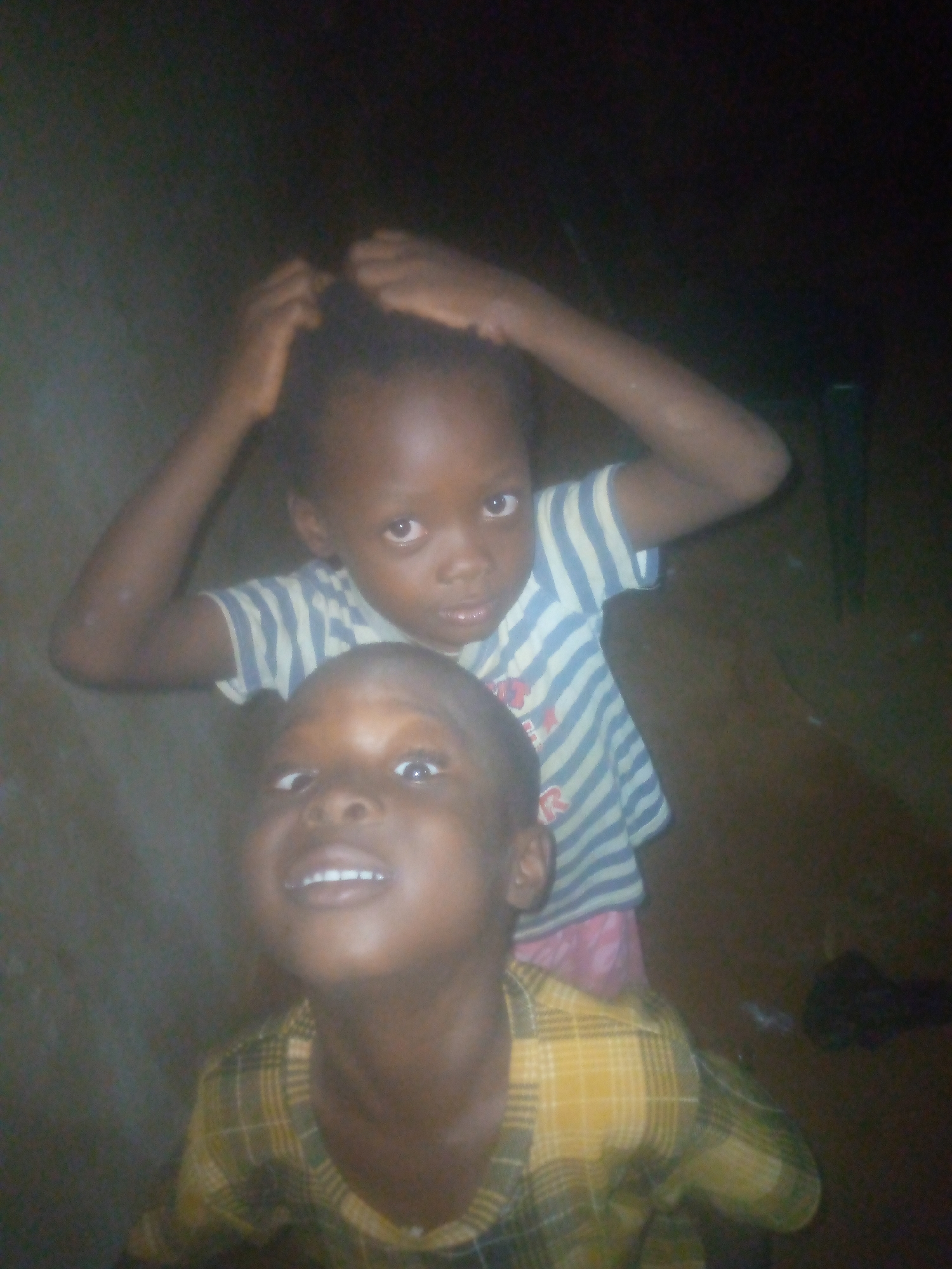 The image displays two young children, appearing to be of African descent, against a dark, indistinct background that suggests a dimly lit indoor space. The lighting is poor, making details hazy and the environment unclear.
In the foreground and lower portion of the frame, one child is visible from the chest up, looking upward with a wide, open-mouthed smile that reveals their upper teeth. This child is wearing a yellow and green plaid collared shirt.
Behind and slightly above, another child is positioned, looking directly towards the camera with a more subdued or curious expression. This child is wearing a blue and white horizontally striped t-shirt. On the front of the t-shirt, a faint red letter "R" is discernible. Their arms are raised, with hands near their head, in what appears to be a playful or self-adjusting gesture.
The time of day is not explicitly clear, but the low light conditions suggest it might be evening or night, or they are in an area with minimal artificial light. There are no visible weather cues or specific outdoor location identifiers. The children's emotions range from joyful (front child) to curious/serious (back child). No other significant objects or text are visible in the scene.