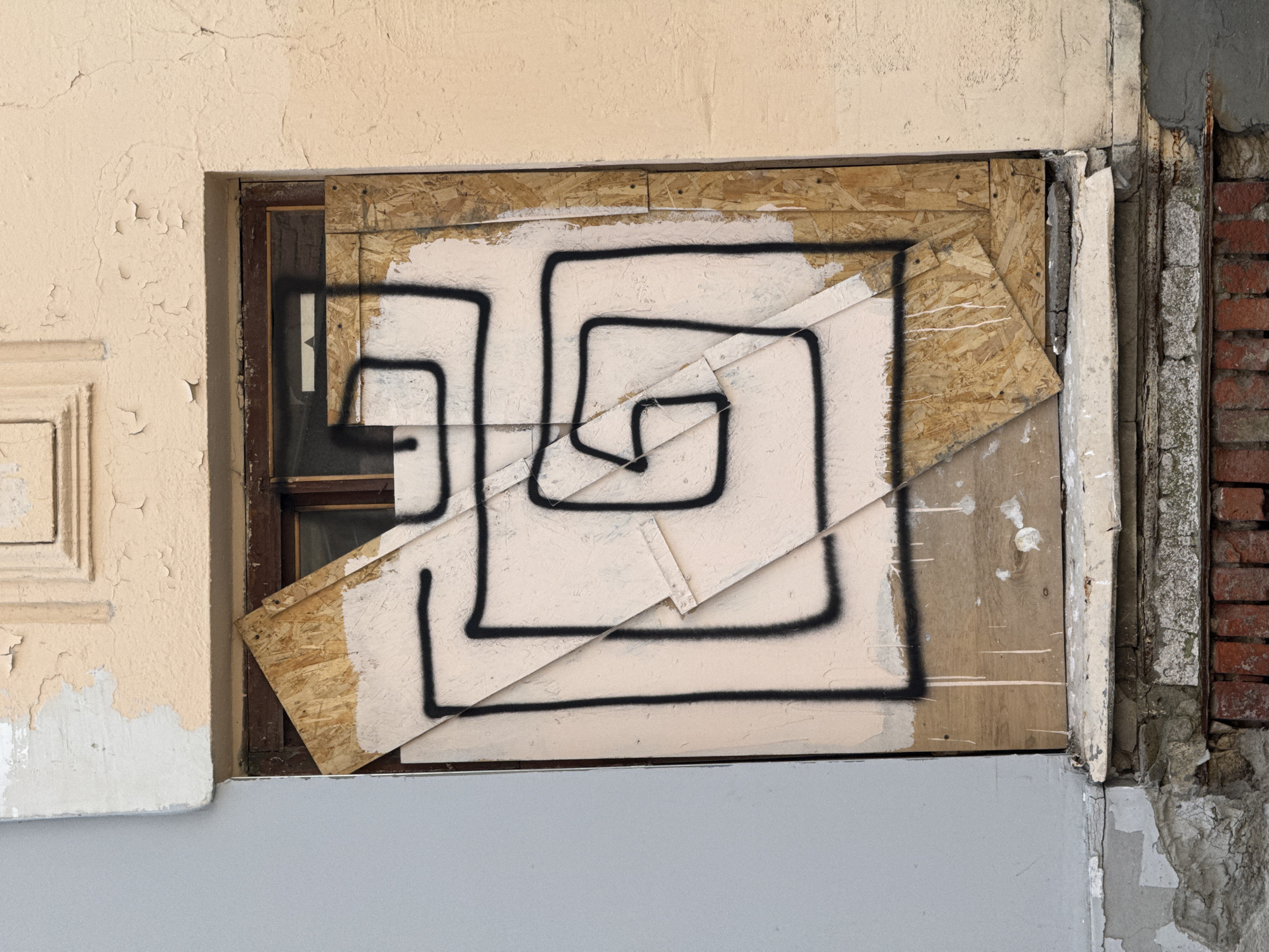 The image shows a boarded-up window in a deteriorated building. The window frame is dark brown. The boards are plywood and painted a light beige with a black, geometric, maze-like design spray-painted on them. The wall to the left of the window is pale yellow with peeling paint and a recessed rectangular design. Below the window is a section of wall painted light gray. To the right of the window, the wall is crumbling, revealing brick underneath. The location is specified as Unknown, Unknown.