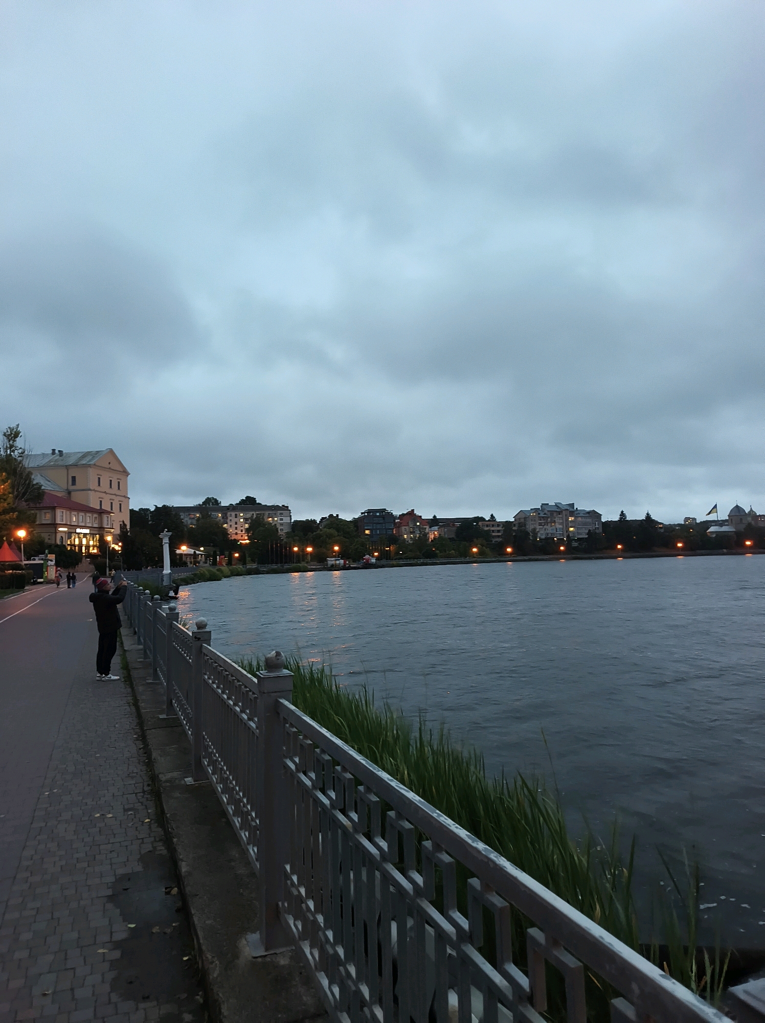 The image depicts a waterfront promenade in Ternopil, Ukraine, during twilight. A person, dressed in dark clothing and a red hat, stands on the paved walkway, holding a phone to take a photograph of the opposite shore. The walkway, made of cobblestones, runs parallel to a metal railing that separates it from the water. Beyond the railing, tall green reeds grow along the water's edge.
Across the Ternopil Pond, a line of buildings is visible. These structures range in height and architectural style, with some featuring illuminated windows. Streetlights with warm yellow lights line the promenade and the distant waterfront, casting a glow on the scene. The sky is overcast with heavy, gray clouds, characteristic of twilight. The water of the pond appears dark and slightly rippled. In the distance, a Ukrainian flag flies on a pole. The overall atmosphere is calm and serene, with a sense of approaching evening.