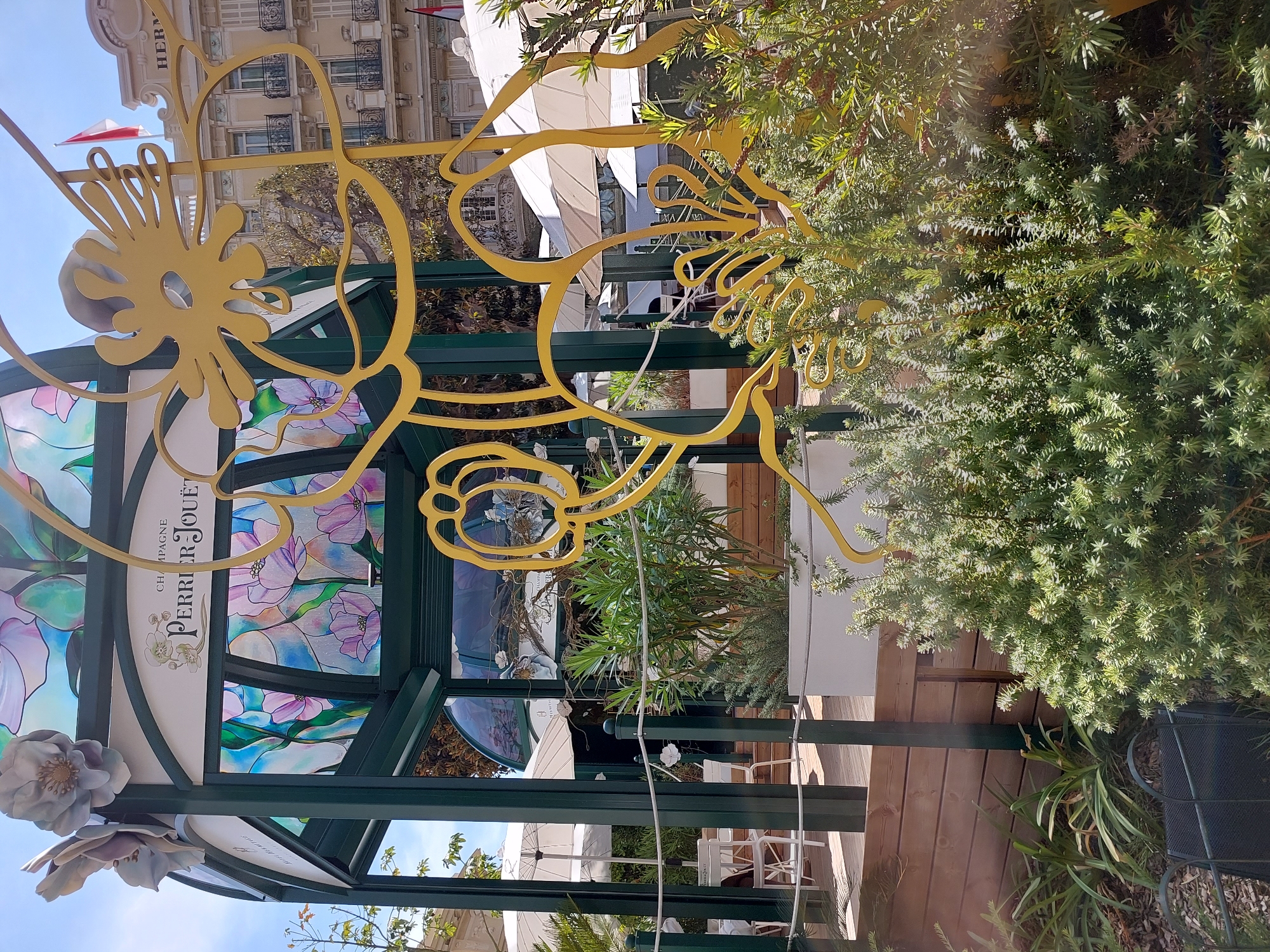 An outdoor promotional kiosk for Champagne Perrier-Jouët is situated in front of a building in Monaco. The kiosk has a green metal frame and features stained-glass panels depicting flowers in shades of purple, pink, blue, and green. A large, yellow, decorative metal sculpture resembling stylized flowers or vines is prominently displayed in front of the kiosk. White outdoor umbrellas and tables with chairs are visible behind the kiosk, suggesting an outdoor dining or seating area. The building in the background appears to be a hotel or apartment complex with classical architectural elements and balconies. A red and white flag, likely the flag of Monaco, is visible atop the building. In the foreground, dense green shrubs and plants partially obscure the view of the kiosk. The scene is set under a bright blue sky.