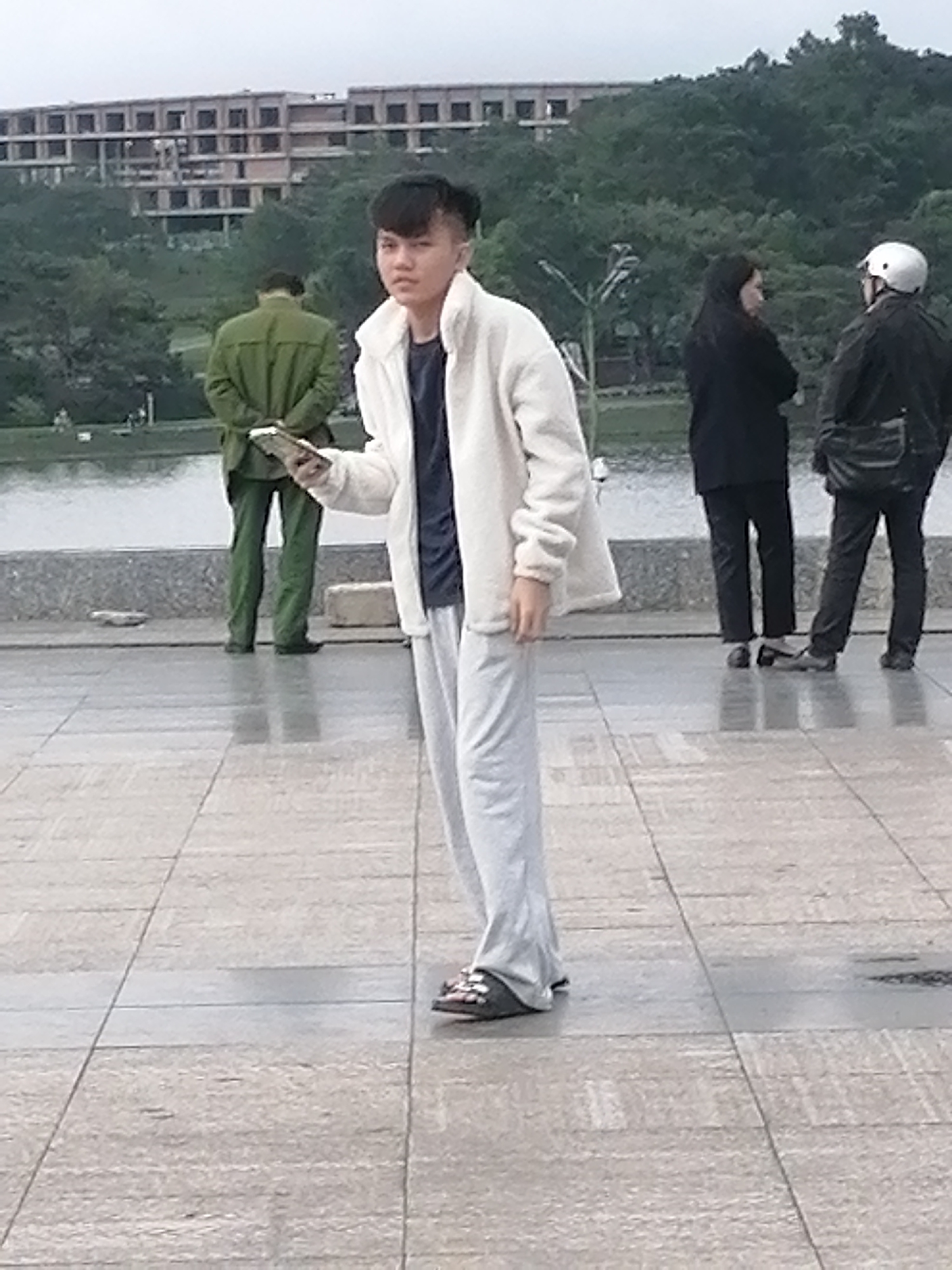 The image depicts an outdoor scene at Xuan Huong Lake in Da Lat, Vietnam. The foreground is dominated by a young person wearing a white fleece jacket, a navy blue t-shirt, and light grey sweatpants. They are holding a smartphone in their left hand and looking towards the camera with a neutral expression. Their footwear consists of dark sandals with white accents.
In the background, to the left, a person in a green uniform, likely a security guard or park ranger, stands with their hands behind their back, facing away from the camera. Further back, on the right side of the frame, two individuals are engaged in conversation near the lake. One person, wearing a black headscarf and black clothing, stands facing towards the lake. Next to them, a person in a black jacket and dark pants, wearing a white helmet, is also facing the lake.
The environment appears to be a paved walkway or promenade. Beyond the walkway is a body of water, Xuan Huong Lake, with trees lining the far bank and a large, partially constructed building visible in the distance. The sky is overcast, suggesting a cloudy day. The ground is wet in places, indicating recent rain.