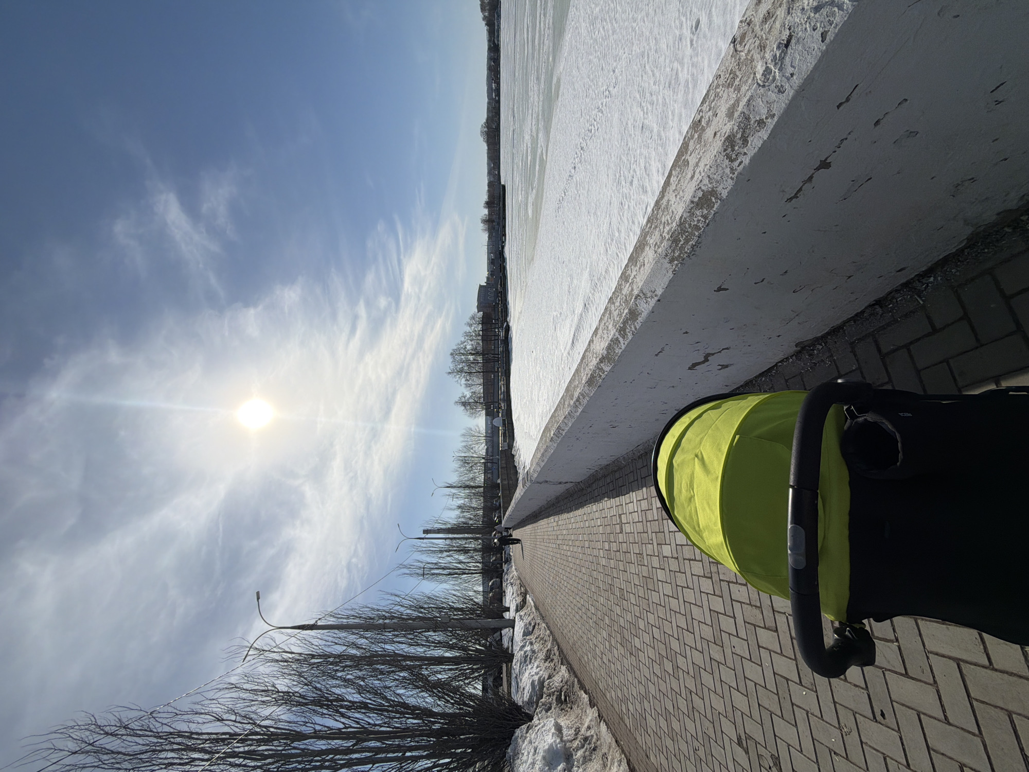 A sunny winter day in Votkinsk, Russia, shows a sidewalk next to a body of water covered in ice and snow. The sky is blue with scattered clouds, and the sun is shining brightly. Bare trees and streetlights line the sidewalk. A person is walking on the sidewalk. A yellow and black stroller is visible. The sidewalk is made of brick.