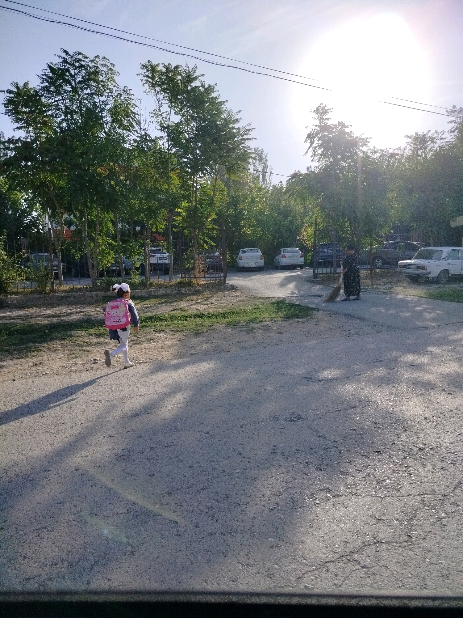 In the city of Kölkent, Kazakhstan, a young girl with a pink backpack runs on a paved road. She wears a dark jacket, white pants, and white shoes, and her hair is tied with a white bow. Further down the road, a woman in a dark, patterned dress sweeps the ground with a broom. Behind them, several cars are parked along the side of the road, and trees line the background. The sun is bright, casting long shadows across the scene.