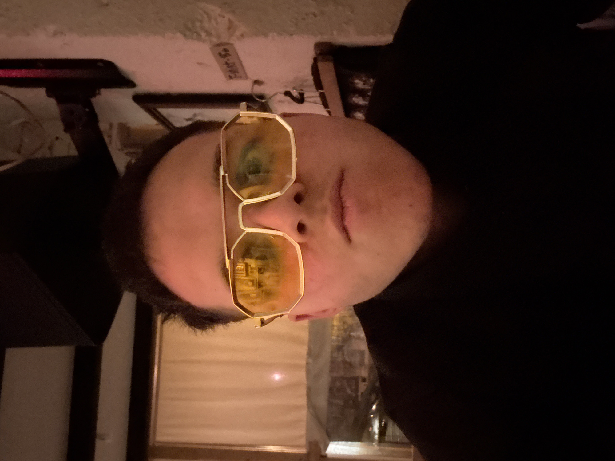A person wearing gold-framed glasses is in an indoor setting. The glasses reflect a room with a lamp and possibly a fireplace. The person is wearing a black garment. The background contains a wall with a small label, a picture frame, and furniture. There is no indication of the city of Sofia, Bulgaria.