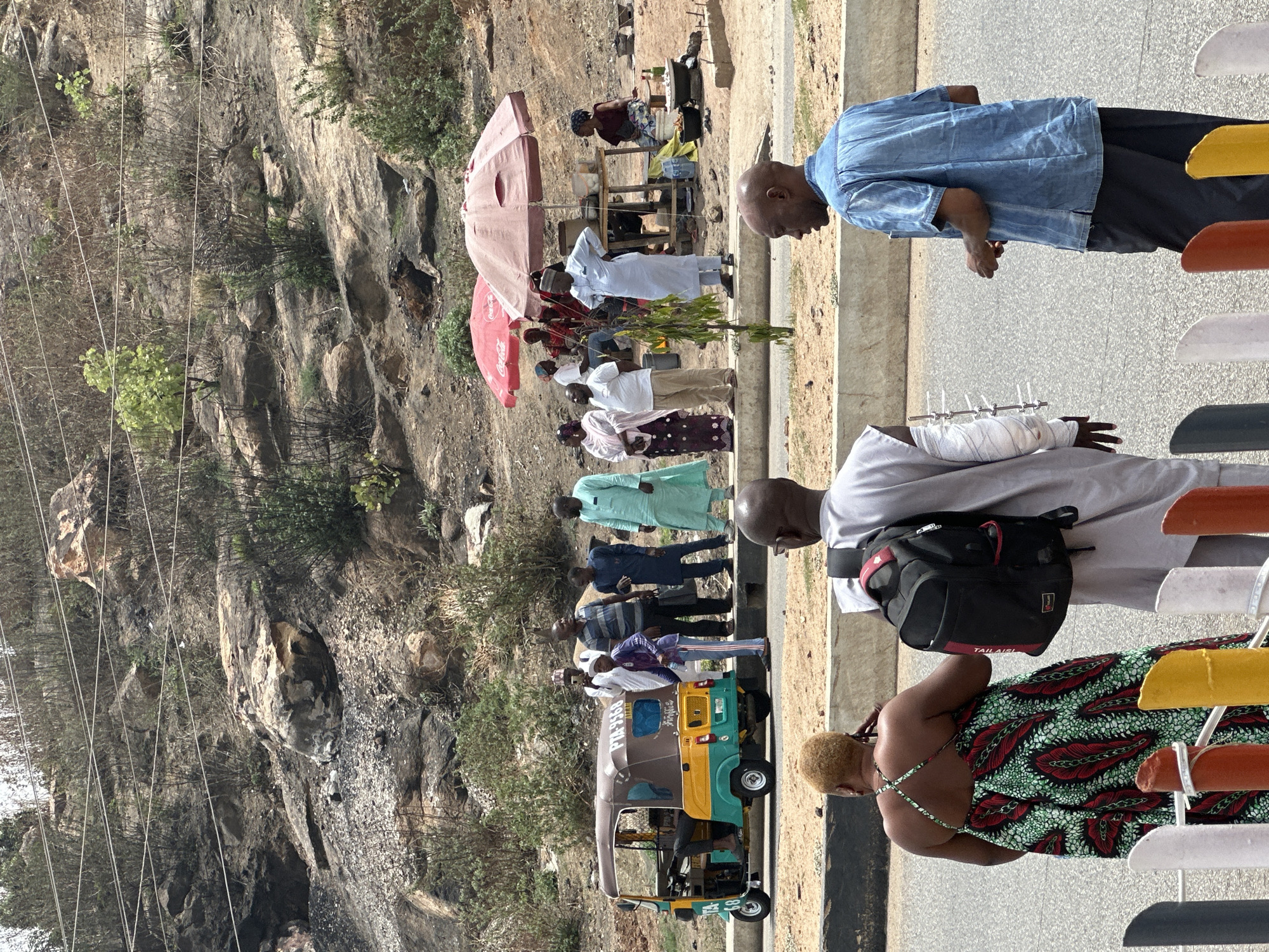 The image depicts a scene along a road in Jos, Nigeria, adjacent to a rocky hillside. A line of people, predominantly dressed in traditional attire, are standing near a green and yellow auto-rickshaw. One man has a bandaged arm with protruding metal supports. Further down the road, there's a small vendor setup under a red Coca-Cola umbrella. Other individuals are scattered around this area. The hillside is rocky and covered with sparse vegetation and overhead electrical wires.