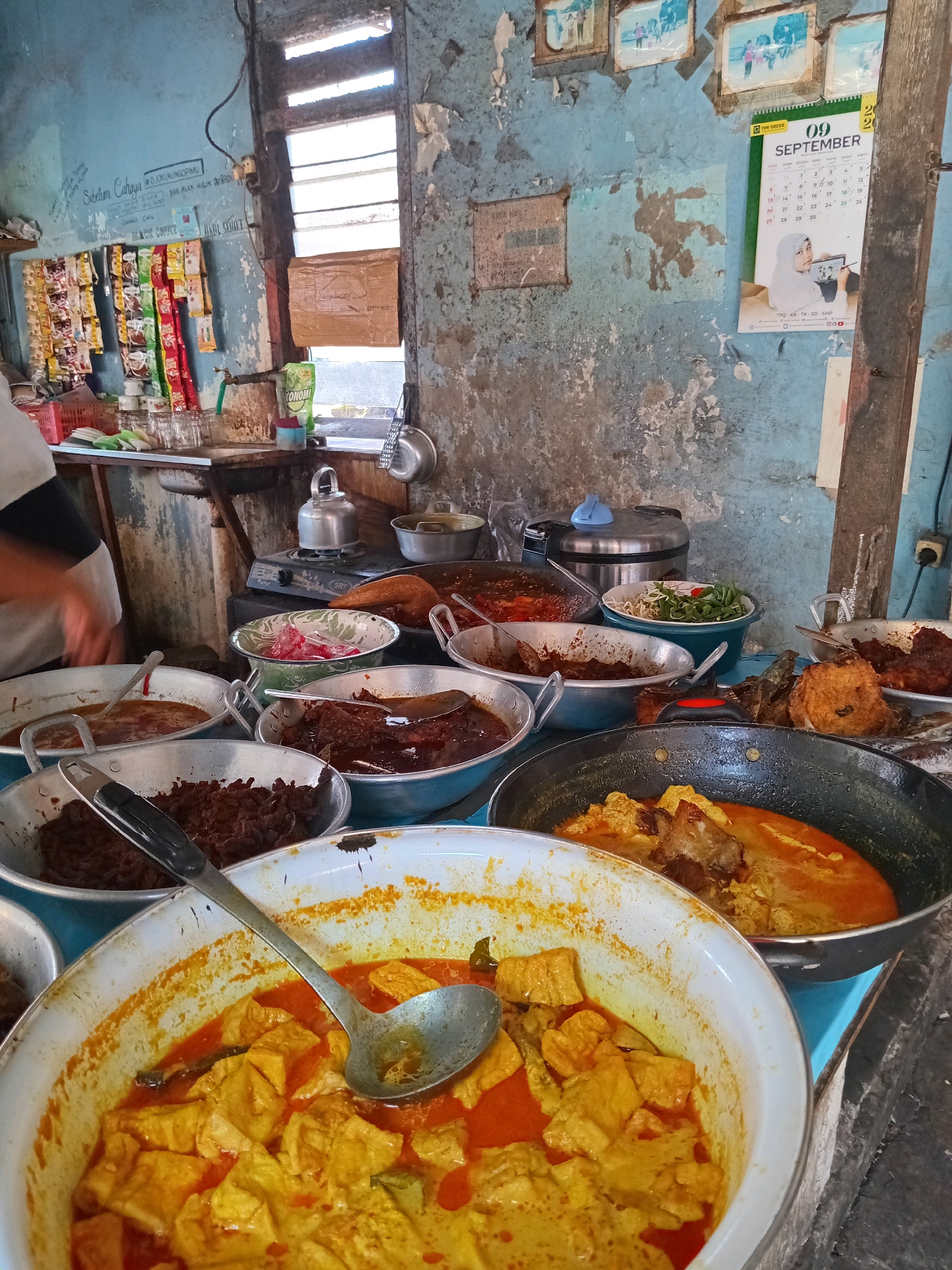 The image depicts an indoor scene in what appears to be a food stall or small eatery, possibly in Ngipik, Indonesia. The environment is rustic, with blue, weathered walls and a rough, textured surface. To the left, a person in white clothing is partially visible, their arm extended as if preparing or serving food.
The central focus is a collection of various dishes displayed in metal bowls and pans. In the foreground, a large white bowl contains a yellow curry with cubed tofu and pieces of meat, with a ladle resting inside. To the right and slightly behind this, a black pan holds a similar curry, with visible chunks of meat. Other bowls contain an assortment of food items, including what looks like shredded meat, a dark stew, and a bowl of vegetables with red chili peppers.
The background reveals more elements of the establishment. Shelves are stocked with packets of instant noodles and other packaged goods. A kettle sits on a small stove. A window with horizontal blinds is visible, letting in natural light. A calendar for September is pinned to the wall, featuring an image of a woman. The overall impression is that of a busy, functional, and perhaps modest food service operation.