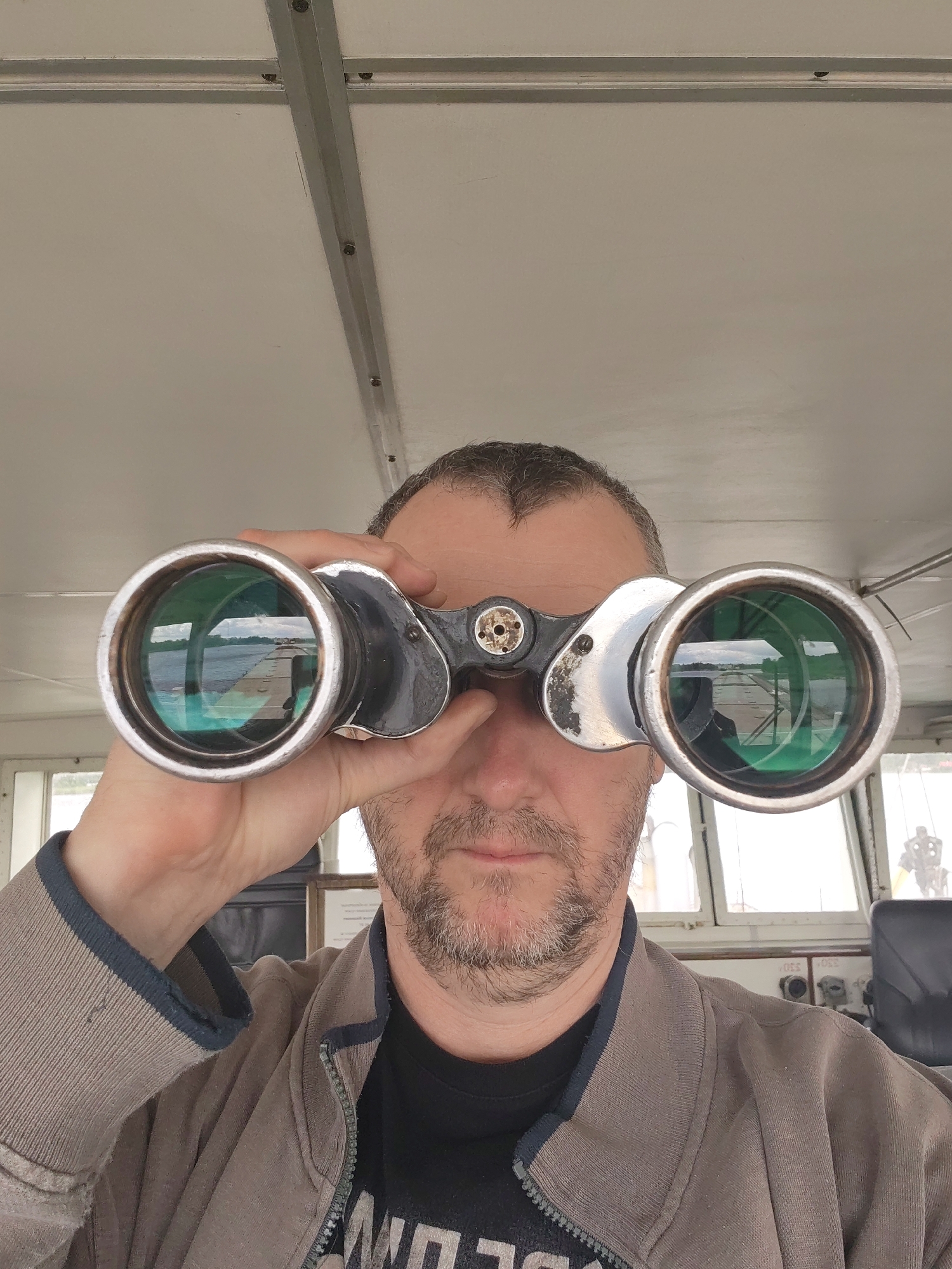 A man holds binoculars to his eyes, looking through them towards what appears to be a pier or dock. The man has short brown hair and a beard. He is wearing a grey jacket over a black t-shirt. The background shows an enclosed space with windows and machinery, potentially a bridge or control room on a boat. The lenses of the binoculars reflect a view of water and a concrete pier, indicating an outdoor setting. Based on the reflection in the binoculars, the scene viewed is consistent with the water features of Белоусово, Russia.