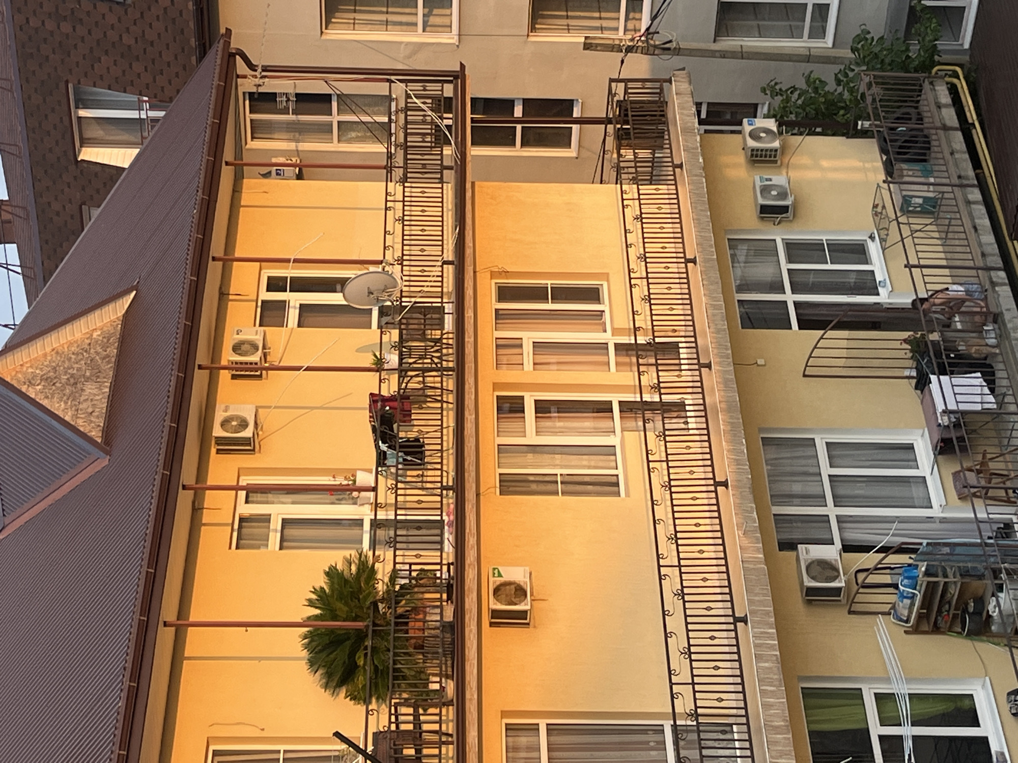 The image depicts a multi-story building in Sochi, Russia, bathed in the warm, golden light of late afternoon sun. The building has a yellow facade and features balconies with ornate black railings on multiple levels. Several windows with white frames are visible, some with curtains drawn. Air conditioning units are mounted on the exterior walls of the building. On the balconies, there are various objects such as potted plants, chairs, tables, and clothes drying on a rack. In the foreground, a portion of a lower balcony shows a shoe rack, laundry, and a person sitting on the balcony. To the right of the main building, another building with a light gray facade and similar windows is partially visible. The background shows rooftops of other buildings, some with brown tiled roofs and dormer windows.