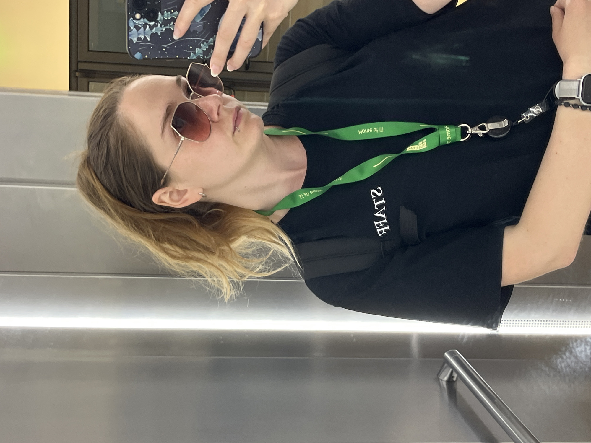 A woman with blonde hair in a ponytail, wearing large sunglasses, a black t-shirt with "STAFF" printed in white, and a green lanyard, takes a selfie in what appears to be an elevator. The elevator walls are metallic and reflective, with a bright vertical light source on the left. Her right hand holds a smartphone with a dark floral-patterned case. A portion of a silver handrail is visible on the left side of the frame. There are no discernible features that place the location specifically in Plovdiv, Bulgaria.