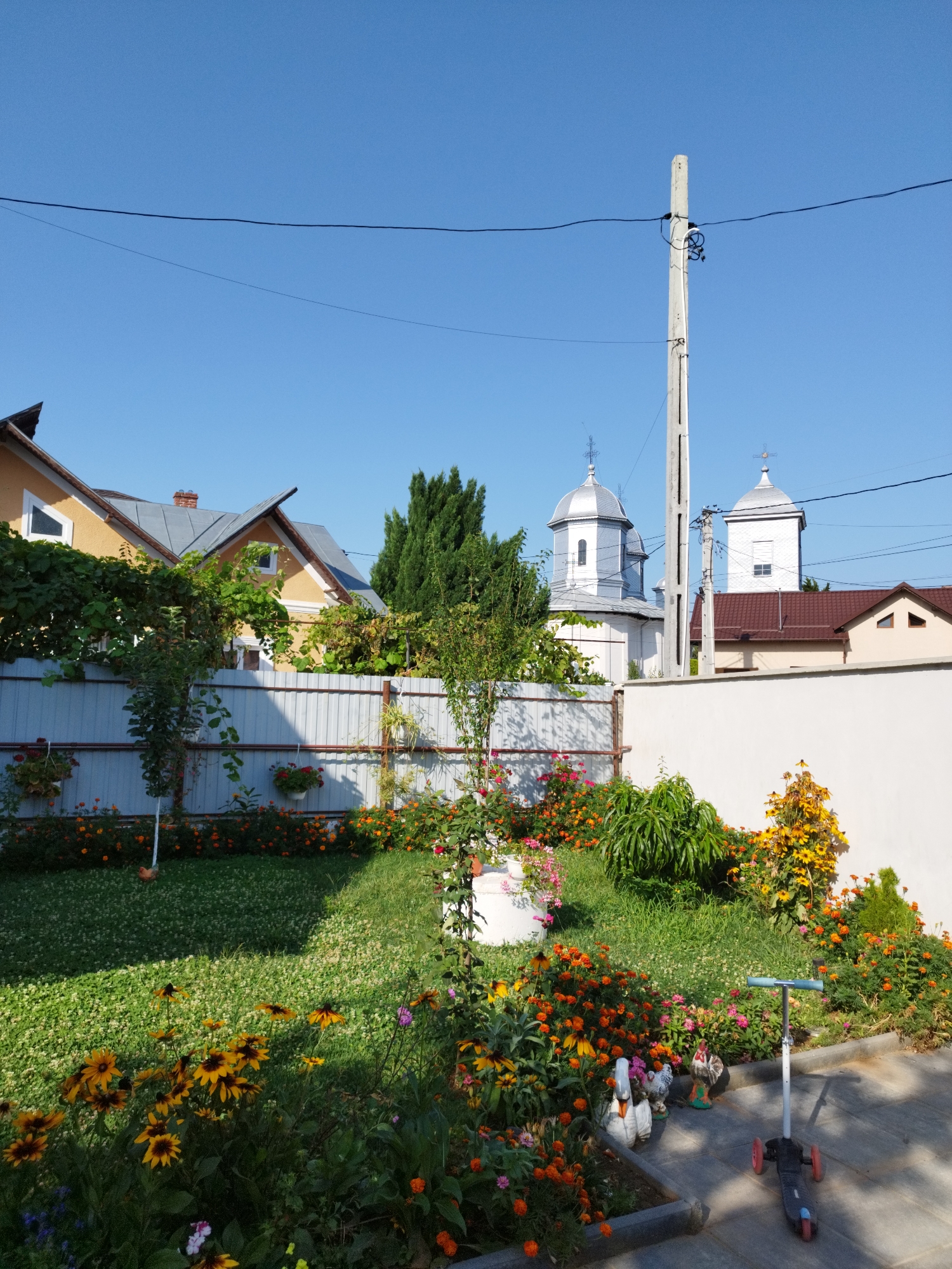 In Băleni-Români, Romania, this outdoor scene captures a sunny day with a clear blue sky. A residential yard is visible, bordered by a light-colored corrugated metal fence and a white wall. Beyond the fence, two church domes with crosses are prominent, suggesting proximity to a place of worship. In the foreground, a lush green lawn is dotted with various colorful flowers, including orange marigolds and yellow coneflowers. A white cylindrical object, possibly a planter, sits in the middle of the lawn. To the right, a gray and blue scooter with three wheels rests on a stone-tiled pathway. Several garden ornaments, including a white swan and two chickens, are placed near the flower beds. The background shows the roofs of houses, with one yellow house in particular being noticeable. A utility pole with electrical wires runs across the sky, adding to the urban or semi-urban setting. The overall impression is one of a well-maintained, cheerful garden.