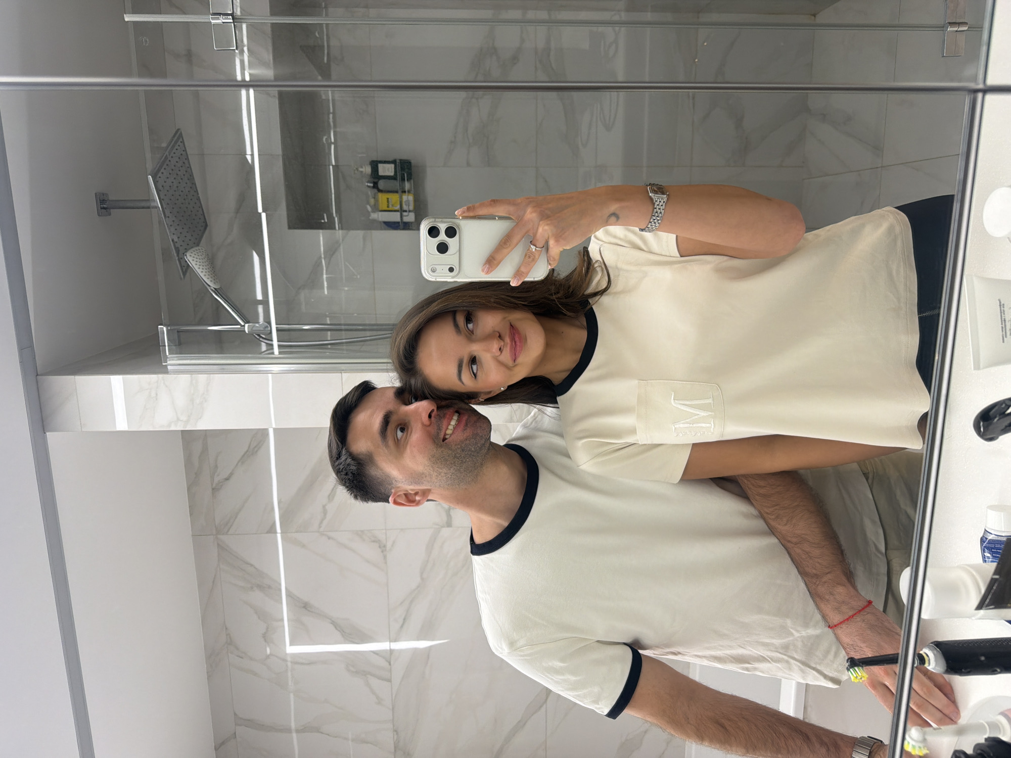 The image shows a couple taking a mirror selfie in a well-lit, modern bathroom located in Krasnoyarsk, Russia. The woman, holding a white iPhone with a clear case, is positioned in front, smiling gently at her reflection. She wears a cream-colored t-shirt with dark trim and an embroidered initial, possibly "M" or "IM," on its chest pocket. She also sports a silver watch and a ring on her left hand, along with a small moon tattoo on her left wrist.
Behind her, the man smiles subtly, looking slightly upwards. He wears a similar cream t-shirt with dark trim on the collar and sleeves and has a red string bracelet on his right wrist. His arm is around the woman, indicating a close embrace.
The bathroom features light-colored marble-patterned tiles on the walls and bright artificial lighting. In the background, a modern shower area with a glass enclosure is visible, showcasing a large, square rainfall showerhead and a handheld shower. On a shelf and counter to the right, various toiletries, including shampoo bottles, toothpaste, and toothbrushes (with brands like "Colgate" and "Oral-B" partially discernible), are neatly arranged. The overall scene suggests a happy, relaxed moment captured by the couple in a contemporary setting.