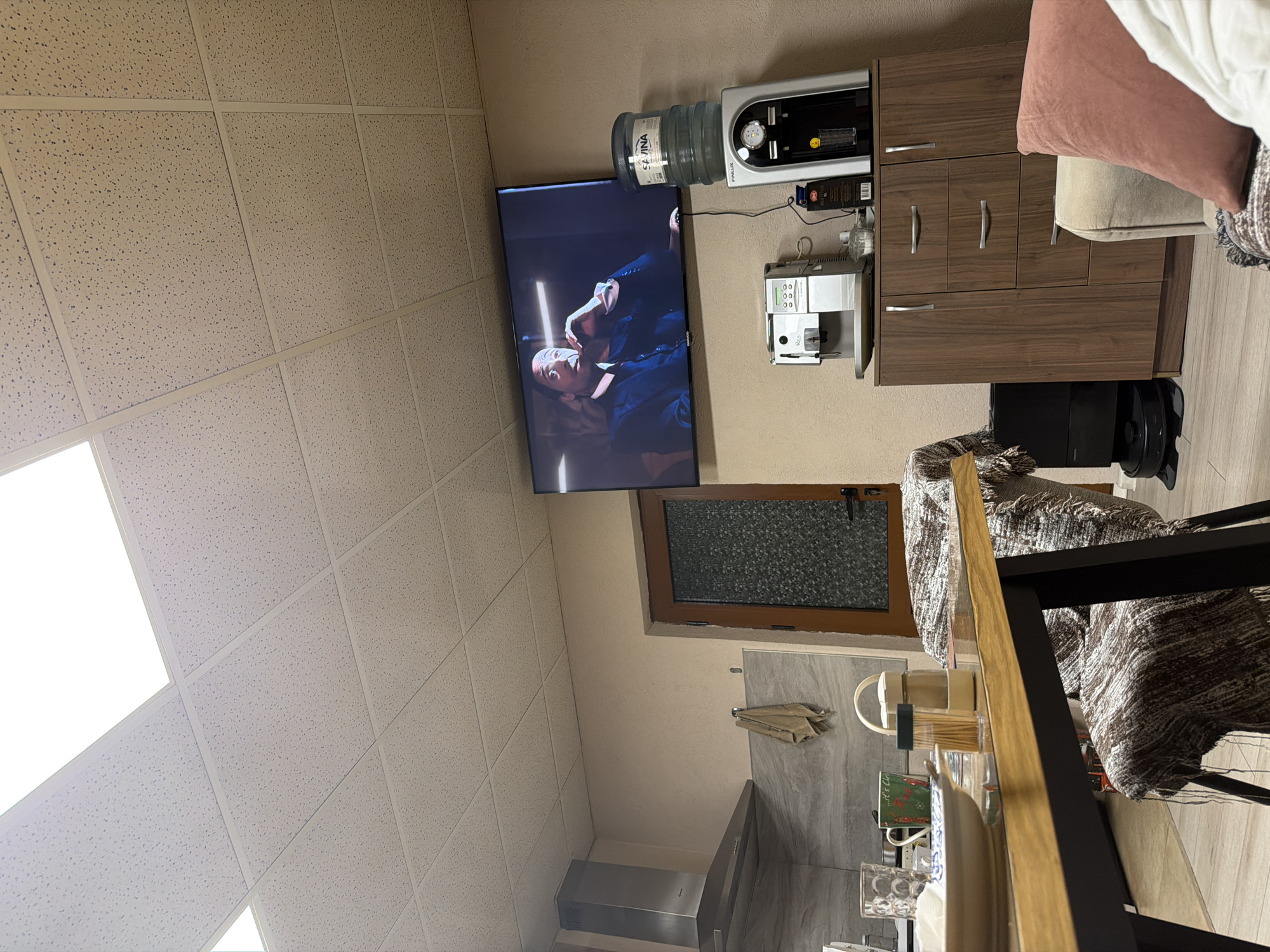 The image shows an interior space with a focus on entertainment and domestic amenities. A television screen displays a scene with people engaged in an ambiguous action. Above it, a water cooler and a coffee maker are mounted on a brown cabinet. Below the TV, a window is visible, and a section of the kitchen with a counter and items such as glassware and a spice rack can be seen. Furniture and accessories, including a cushion and a woven throw, suggest a comfortable living area.
There is no information in the image to identify the location in the city of Panagyurishte, Bulgaria.