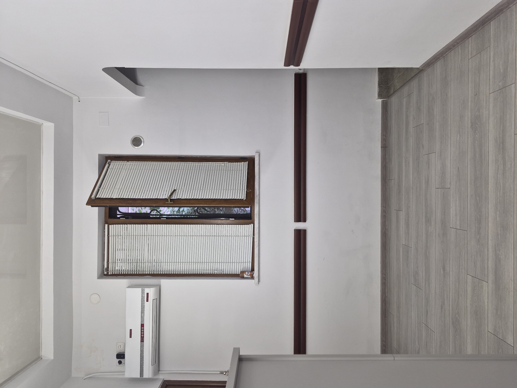 The image depicts an interior hallway or room in Yerevan, Armenia, characterized by stark white walls and a light-colored wooden plank floor. Dominating the central wall is a wooden-framed window with white horizontal blinds, partially open to reveal greenery outside. To the left of the window, a white air conditioning unit is mounted high on the wall. Below the window and along the walls, dark brown wooden horizontal panels are installed as a wainscoting or protective barrier. The ceiling is white and appears to have a recessed lighting fixture. A doorway or archway is visible on the right side, leading to another area. The overall impression is one of a clean, modern, and functional space, possibly a reception area or a common corridor in a building.