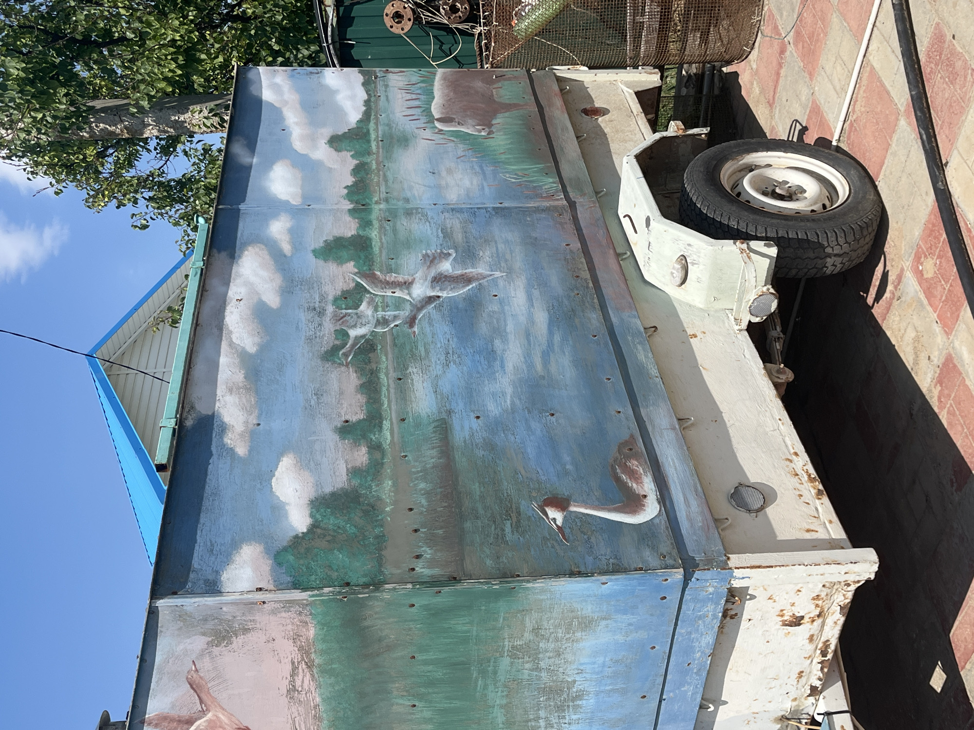 The image shows a weathered, white trailer with a large, rectangular metal cargo box painted with a mural of a natural landscape. The mural depicts a body of water, trees, clouds, birds in flight, a bird with a red crest in the water, and a wild boar standing in reeds. The trailer itself is white, with visible rust and peeling paint, particularly on the lower sections. It has one visible wheel with a black tire and a white rim. Behind the trailer, a blue-roofed building is partially visible, along with trees and a large wooden pole. To the right of the trailer, a rusty metal cage and some industrial-looking metal objects are present. The ground is paved with red and white square tiles. The overall impression is one of an outdoor storage area, possibly in a rural or semi-rural setting within Krasnyye Barrikady, Russia, given the presence of a mural featuring local wildlife and the general aesthetic.