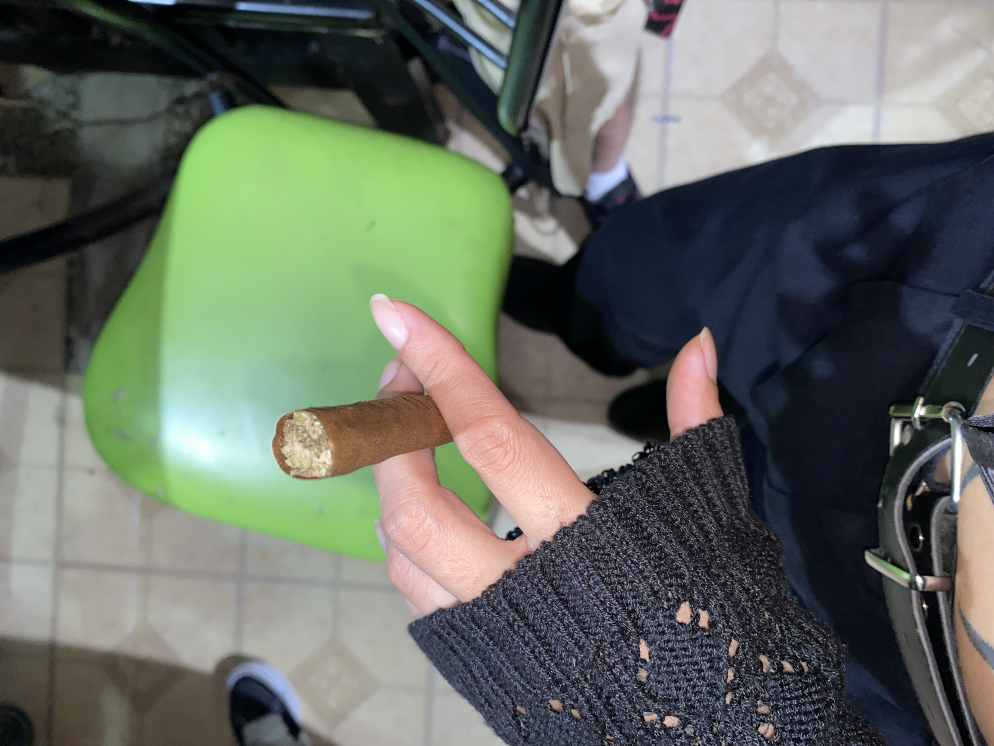A high-angle shot depicts a hand holding a lit cigar, situated above a green plastic chair, with a portion of a black garment and belt visible in the background. The location is possibly Monte Grande, Argentina. The hand is adorned with a black, open-weave sleeve. The cigar's lit end is held between the thumb and index finger. The background consists of tiled flooring and glimpses of a black shoe. The scene suggests an individual seated on a chair, smoking a cigar in an indoor environment.