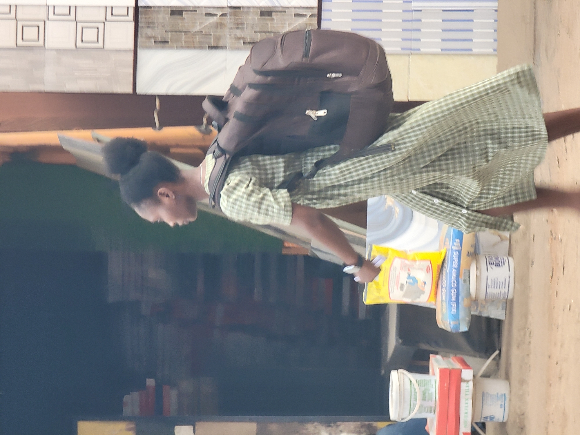 A woman stands in front of a storefront that displays tiles, with a brown backpack. She wears a green checkered shirt and has her hair in a bun. She is holding a variety of construction supplies. Among these supplies is a bag labeled "SUPER AMACO GUM POW," as well as pails and other construction materials. A stack of red tiles is visible. The scene is potentially in Maiburiji, Nigeria, as construction supplies are typically sold in commercial districts.