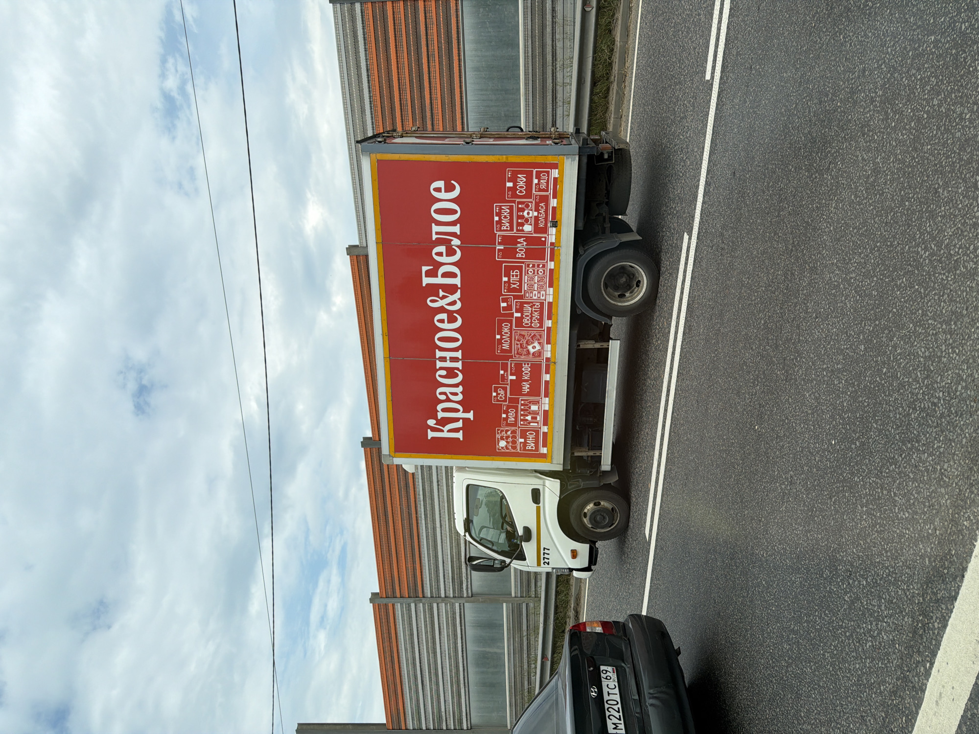 A white truck with a red trailer is parked on a road in Radumlya, Russia. The trailer has "Красное&Белое" written in large white letters. Below the name, there are smaller images and text indicating various products such as milk, bread, wine, and juices. The truck has the number "2777" on its side. A black car is visible to the left of the truck. In the background, there is a sound barrier along the side of the road. The sky is cloudy.